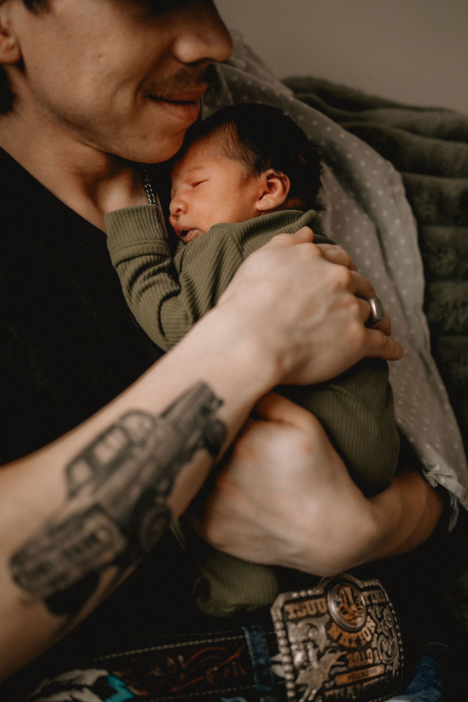 Tattooed Calgary parent tenderly holding sleeping newborn baby dressed in green, sharing a gentle moment.