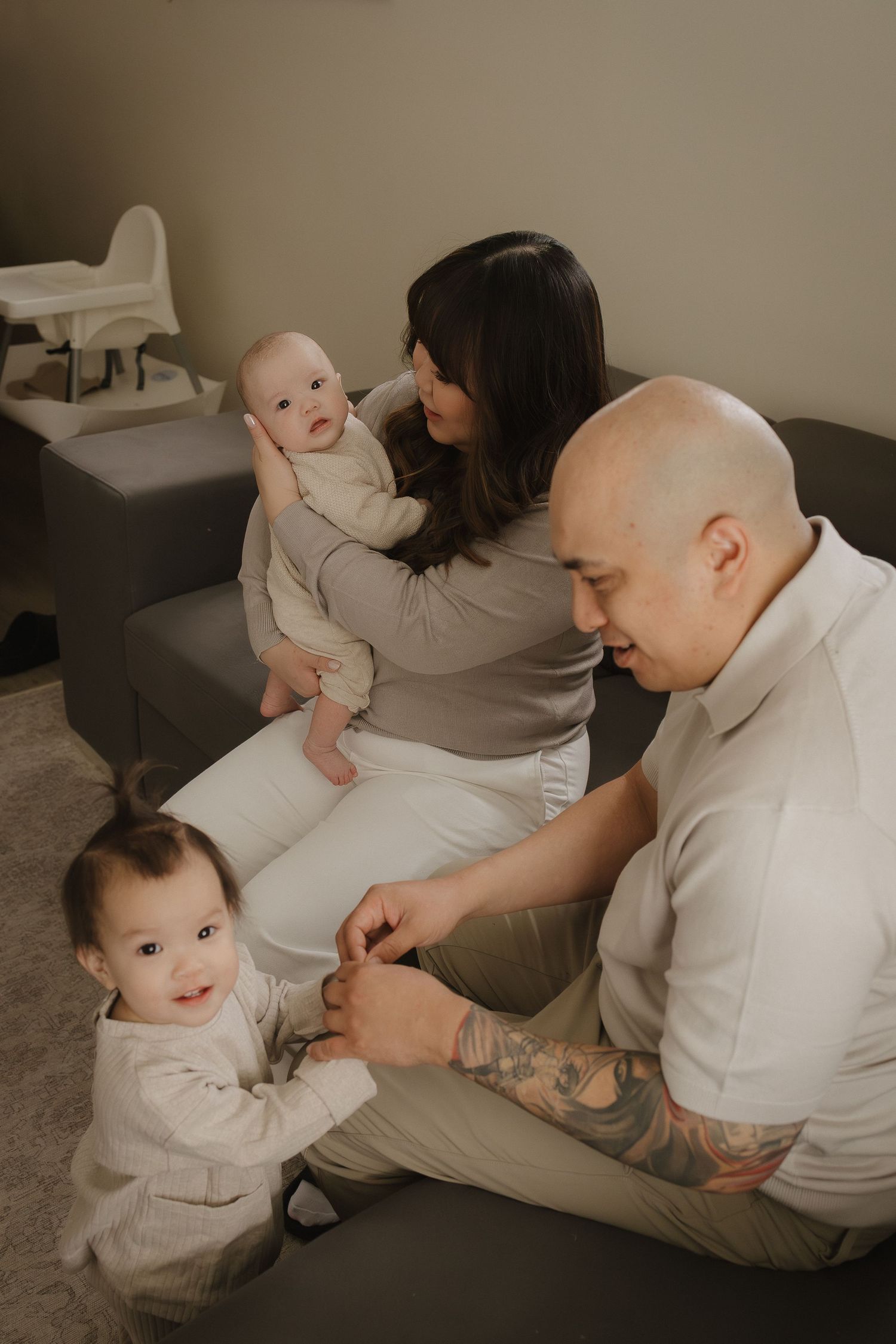 A young family with two babies sits on a couch, dad with tattoos, mom holding an infant for a Calgary in-home photography session.