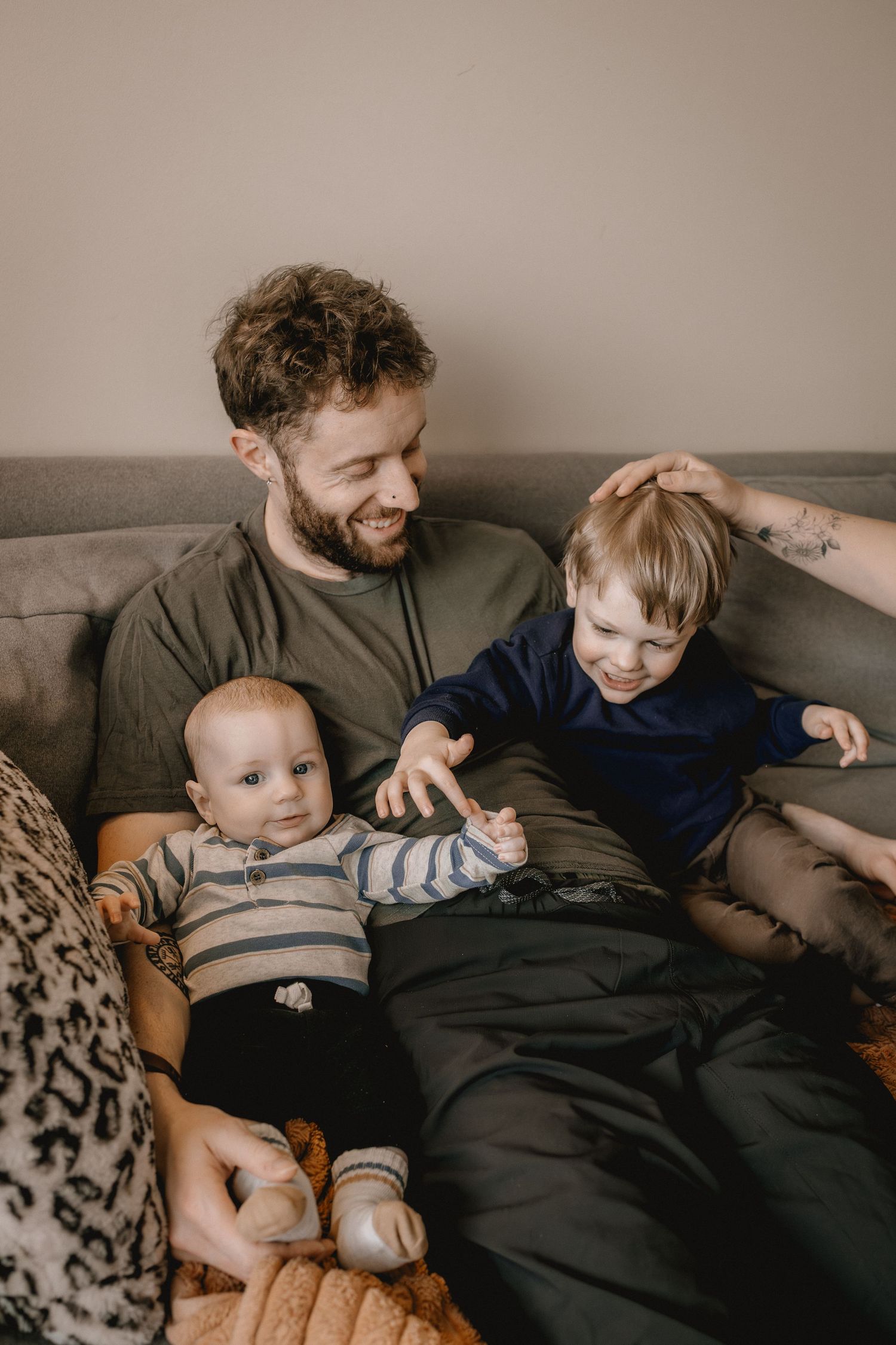 Smiling bearded father sitting on couch with infant and toddler sons, enjoying a warm family moment at family photography session