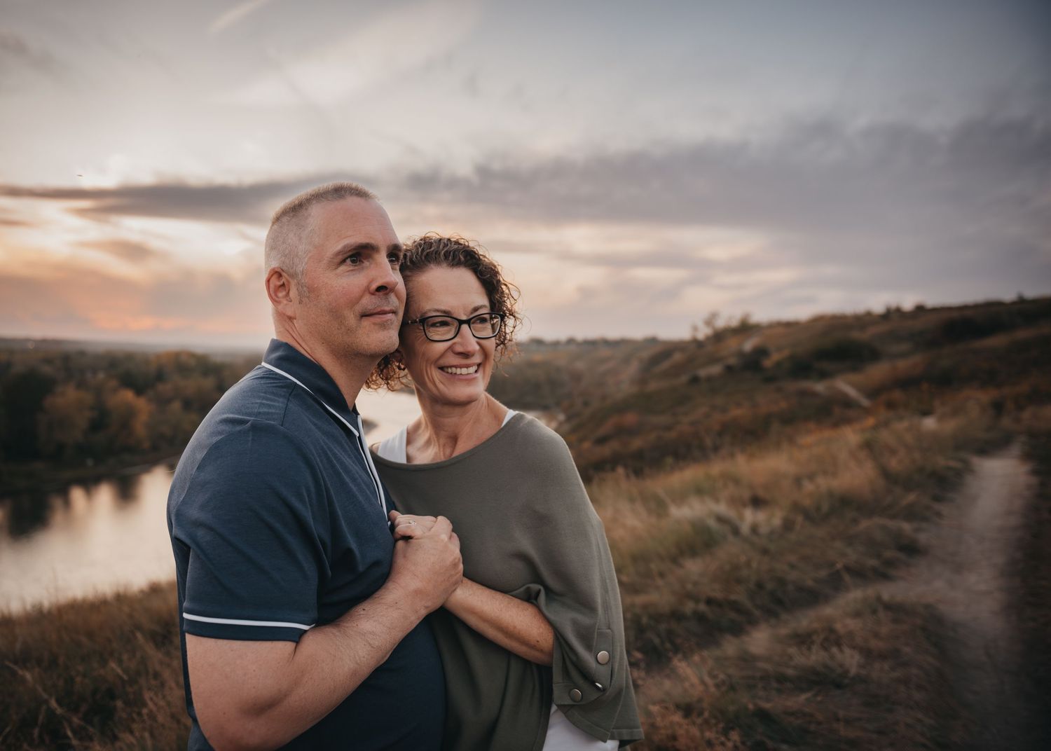 Couple embracing outdoors at sunset near a river, smiling woman in glasses hugging man in navy polo shirt.