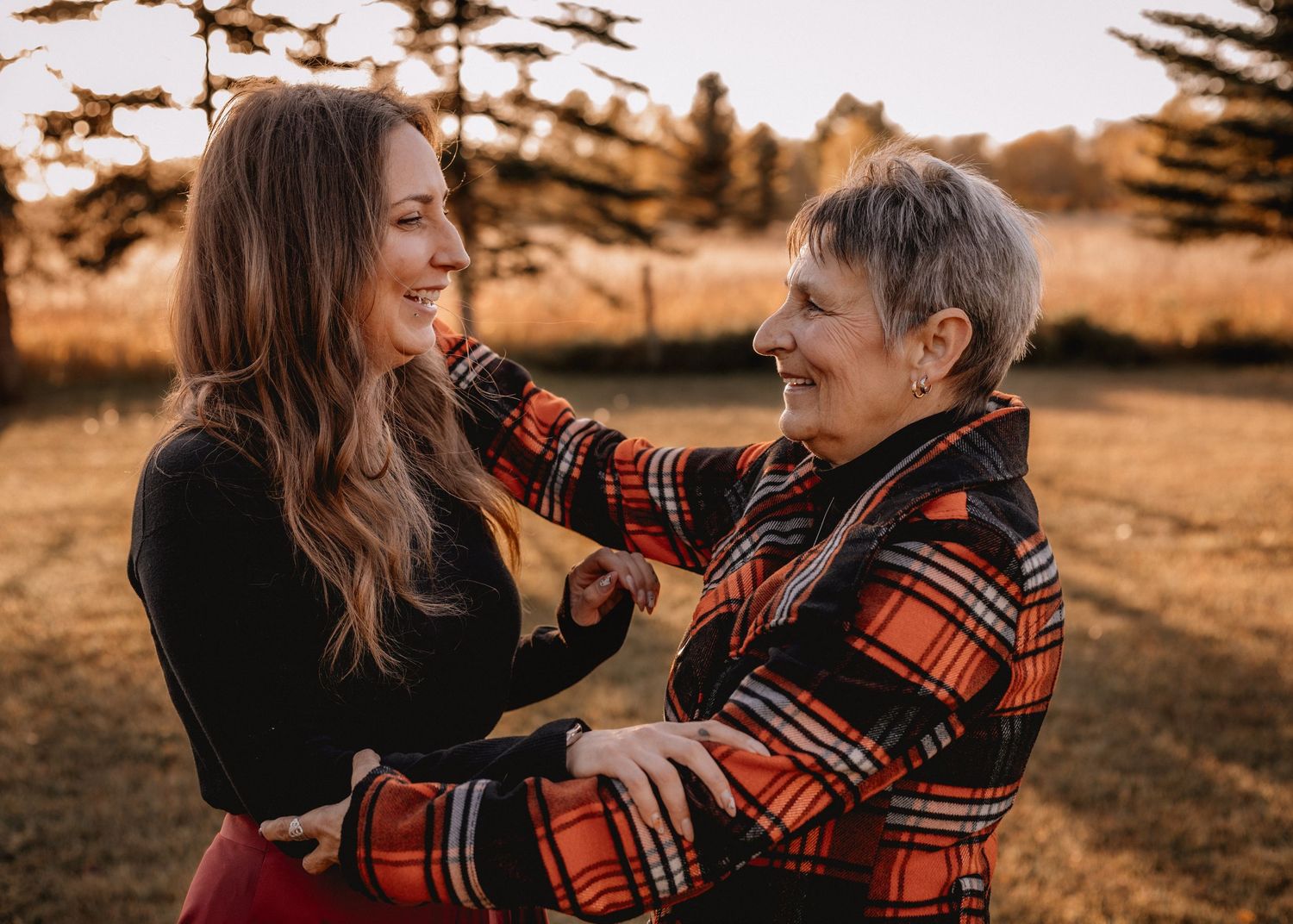 Two women laughing and embracing outdoors in a golden High River field, wearing plaid and black outfits during autumn.