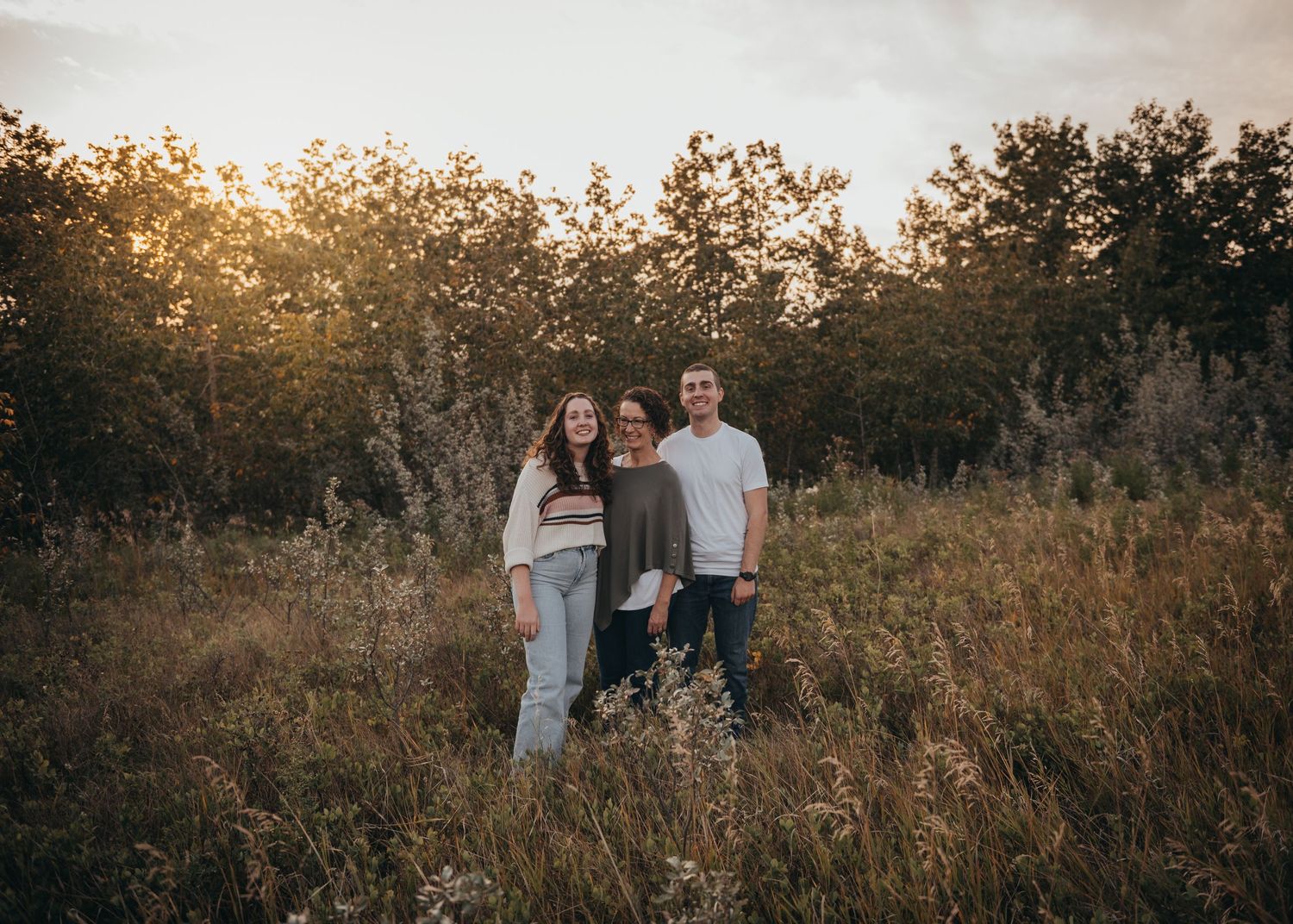 Three people smiling together in a golden sunlit Calgary field surrounded by tall grass and trees at sunset.