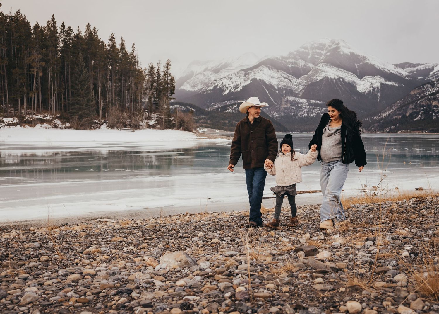Family of three walking along rocky lakeshore with snowcapped mountains and frozen Barrier Lake in winter landscape during maternity session