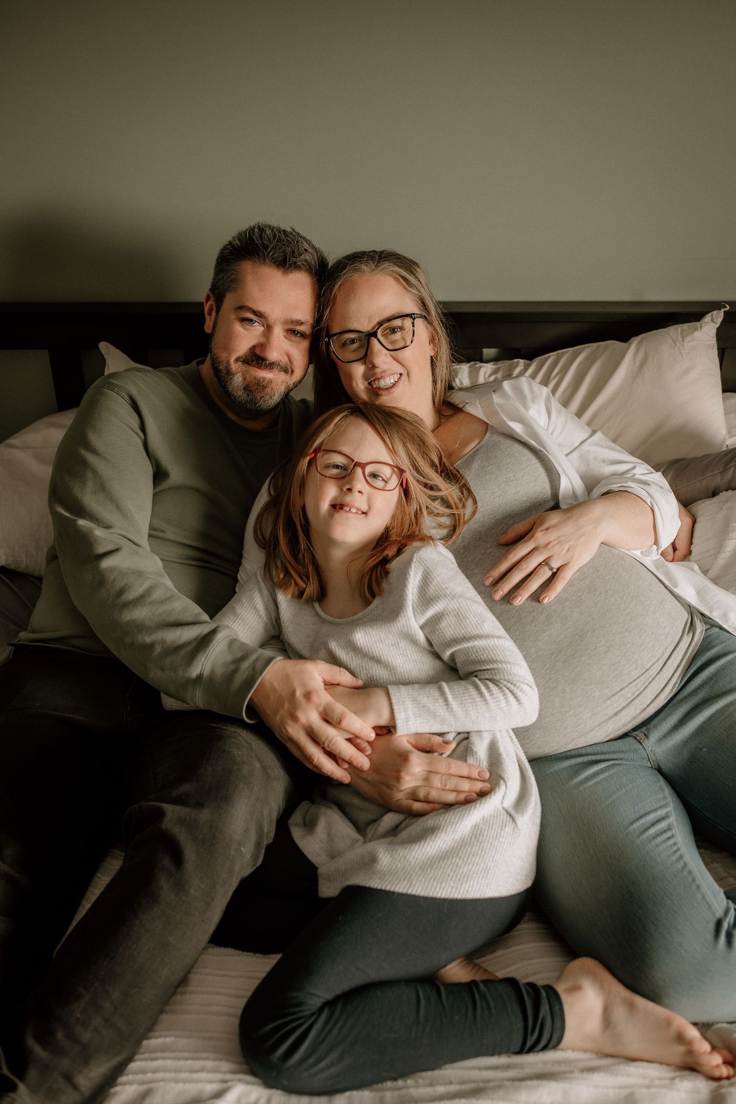 Candid photo of a happy family of three on a bed, with a pregnant mother, father, and young girl with glasses hugging her mom's belly.