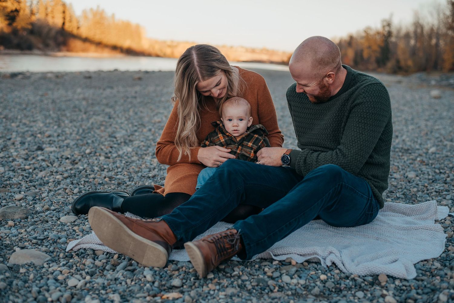 Family of three sitting on rocky beach at Bowness Park, parents admiring their baby on a white blanket during golden hour.