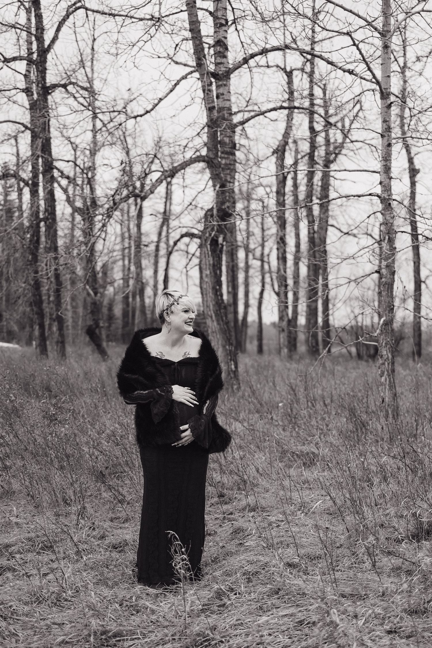 Pregnant woman in black dress and fur wrap poses among bare trees in a moody black and white outdoor portrait.