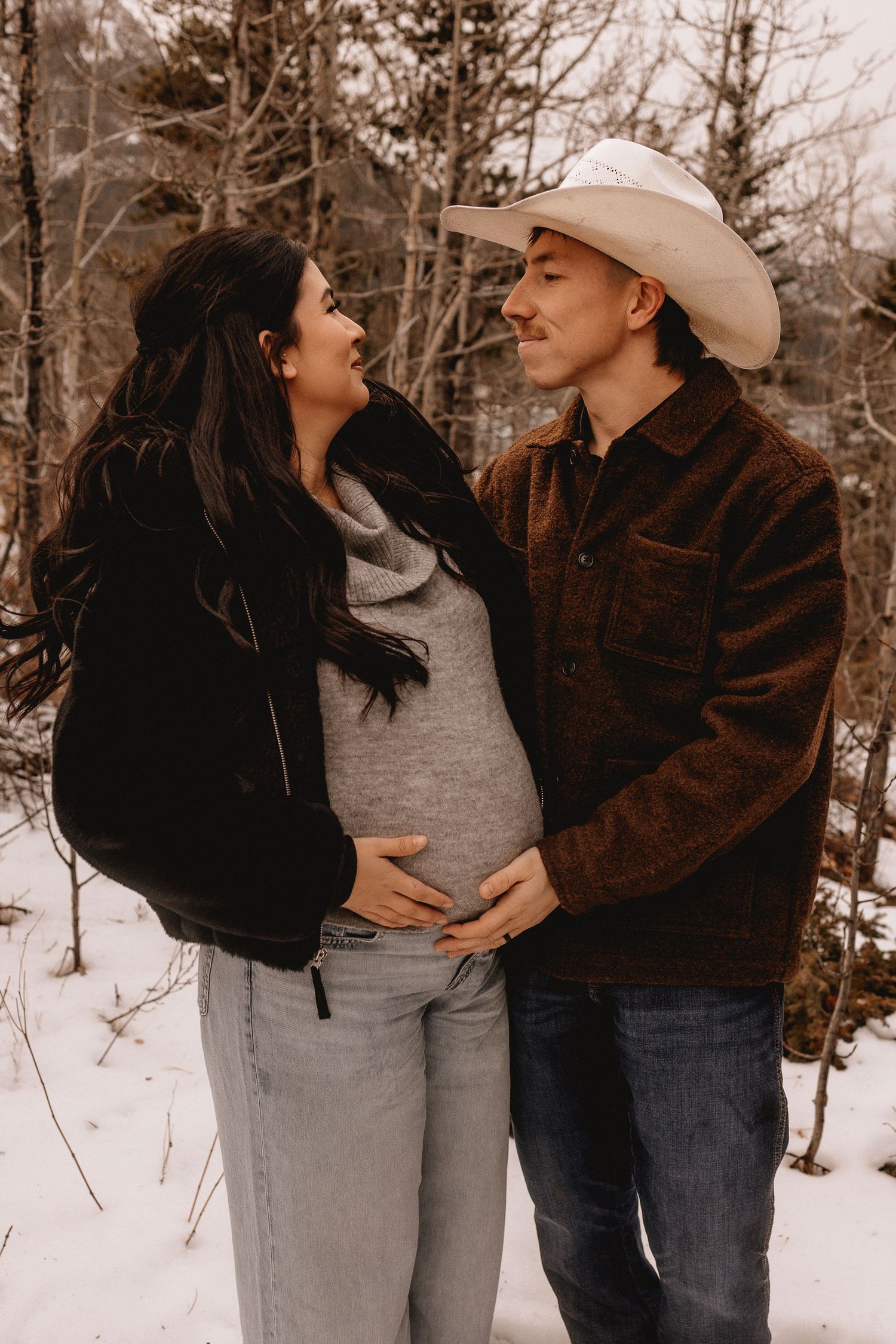 A Calgary couple shares tender moments in a snowy winter forest setting wearing casual clothing and a cowboy hat.