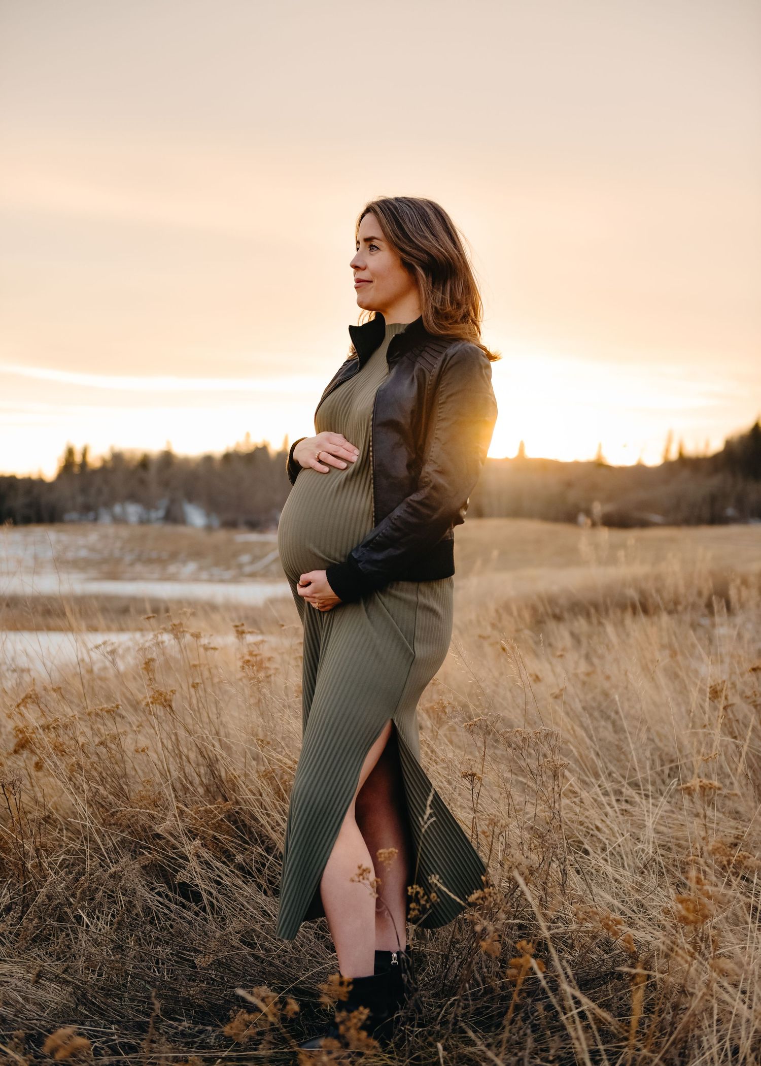 Pregnant woman in olive dress and leather jacket standing in golden Calgary field at sunset, cradling baby bump.
