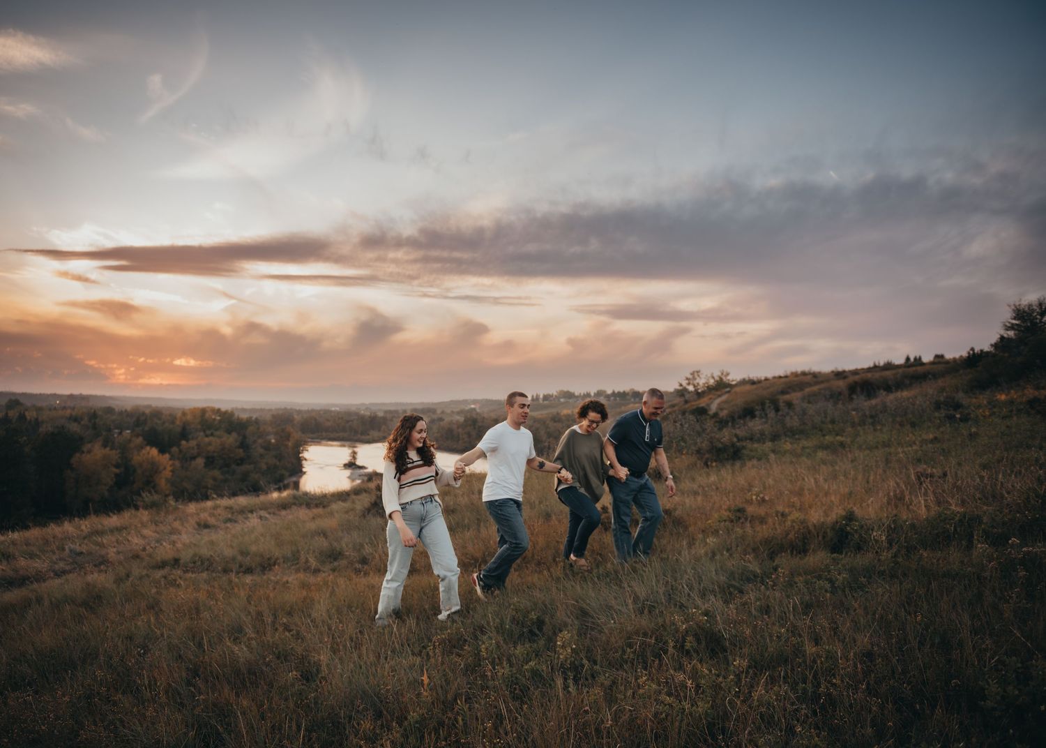Four people walk hand-in-hand through a grassy Calgary field at sunset with rolling hills and a river in the background.
