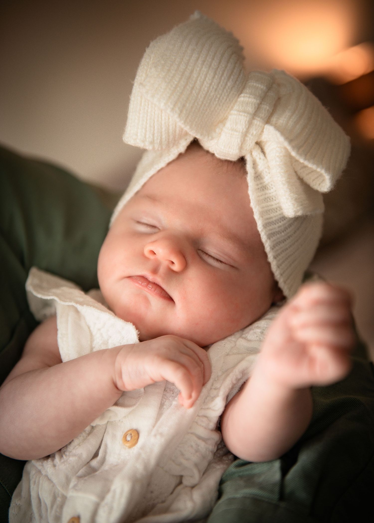 Sleeping newborn baby wearing a cream knit bow headband and white outfit, peacefully resting with tiny hands raised.
