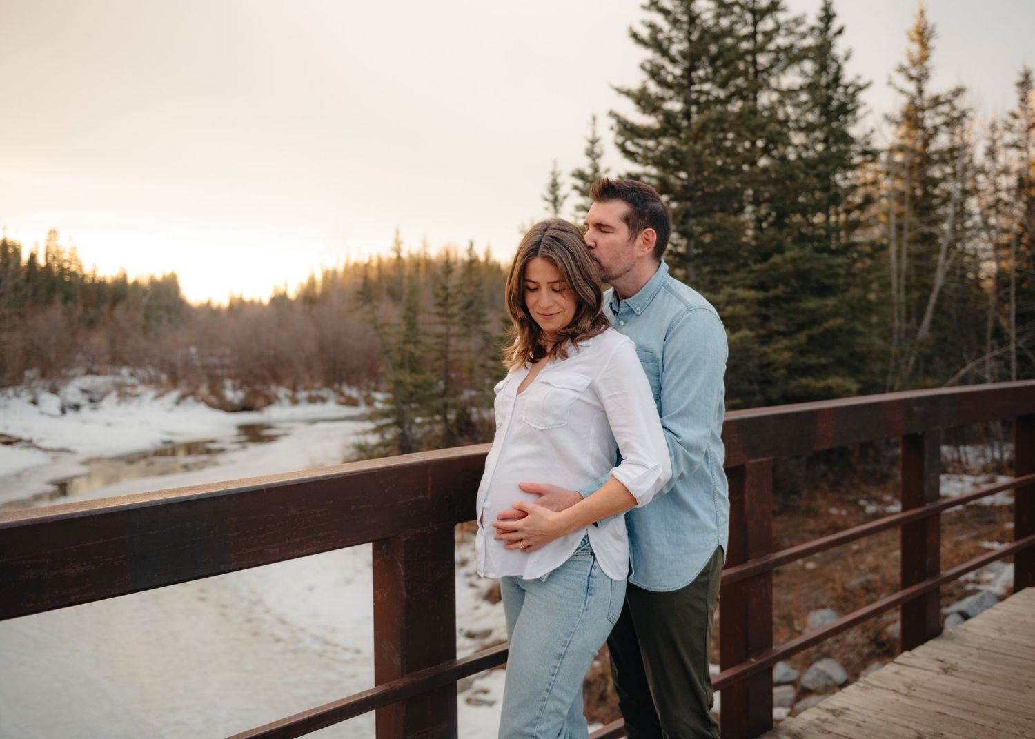 A couple shares an intimate moment on a wooden bridge in Calgary, AB overlooking a snowy winter landscape with evergreen trees.