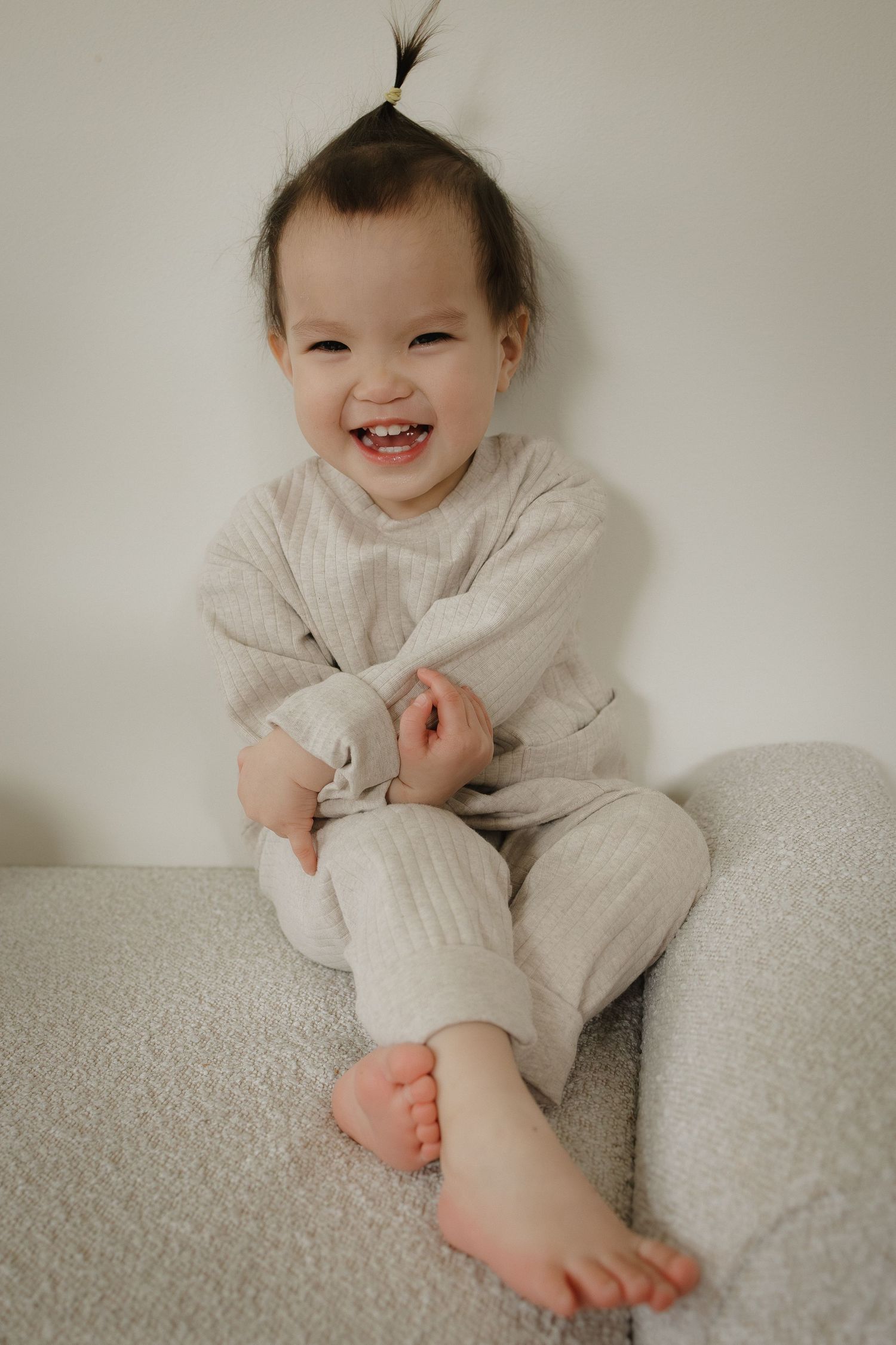 Smiling toddler girl in beige pants sitting barefoot on a soft gray surface against a white wall posing candidly for a Calgary photographer.