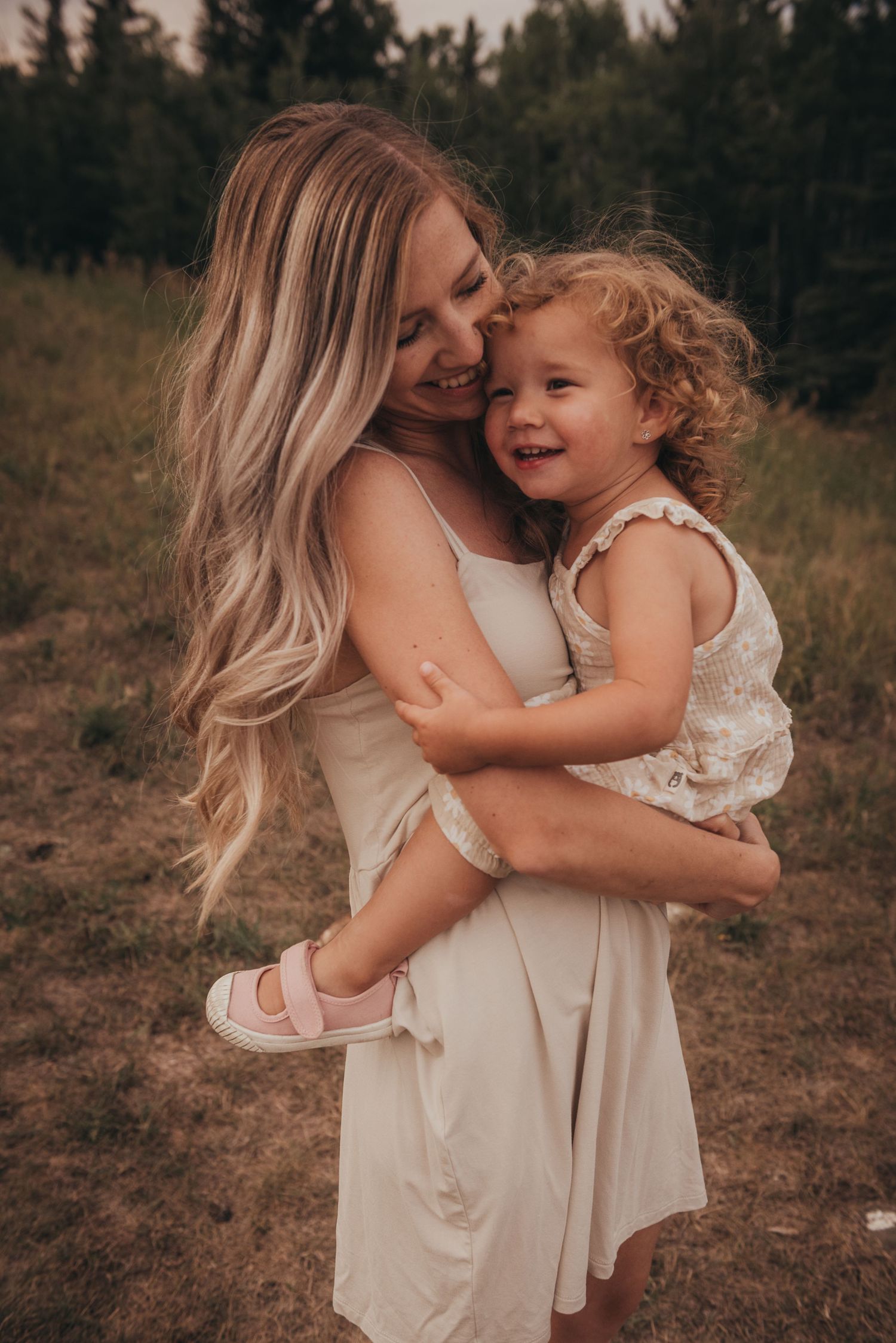 A mother with long blonde hair in a white dress lovingly holds her smiling toddler daughter outdoors during family photoshoot.