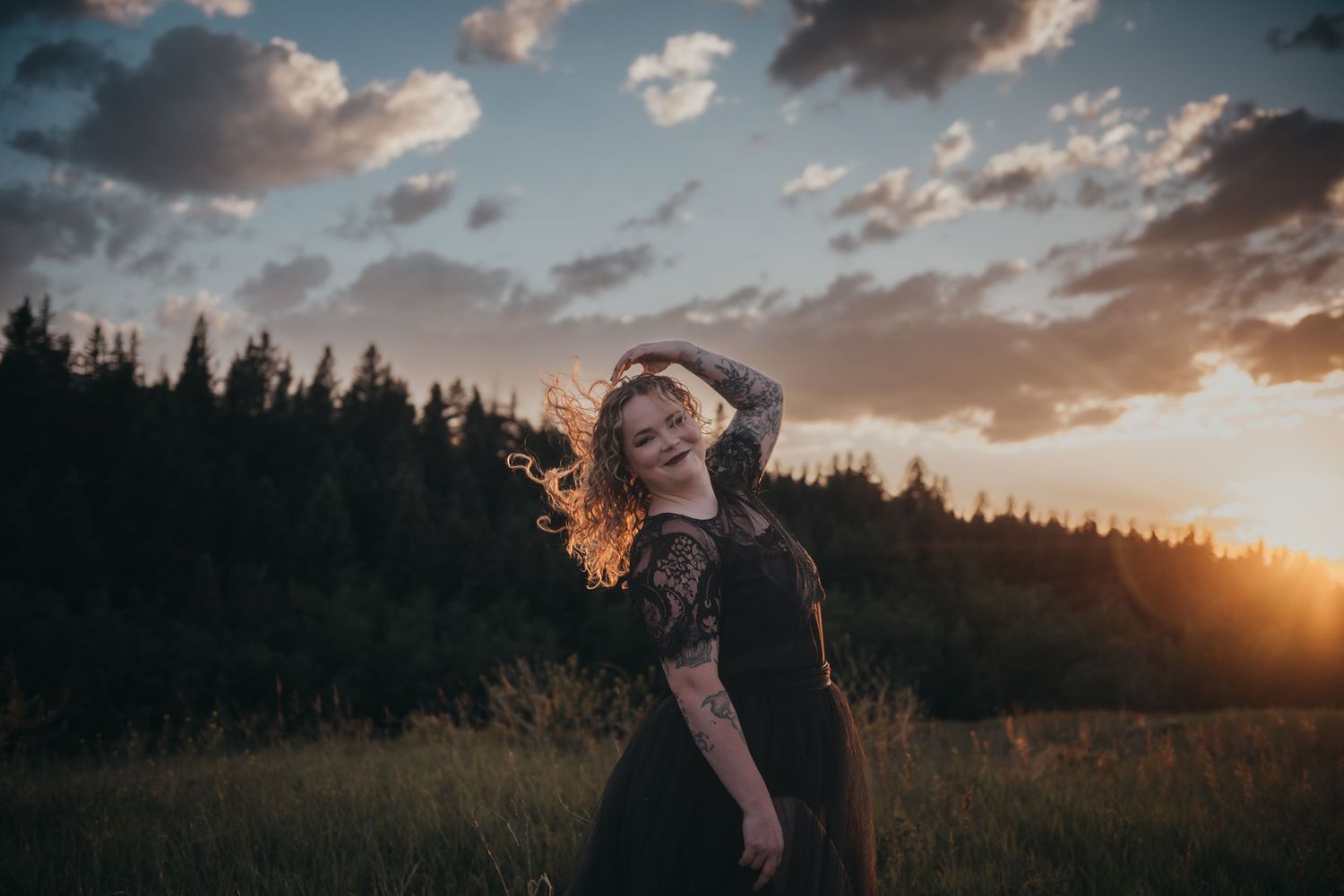 A young woman in a black lace dress dances in a Calgary, AB meadow at golden sunset with forest and dramatic clouds behind her.