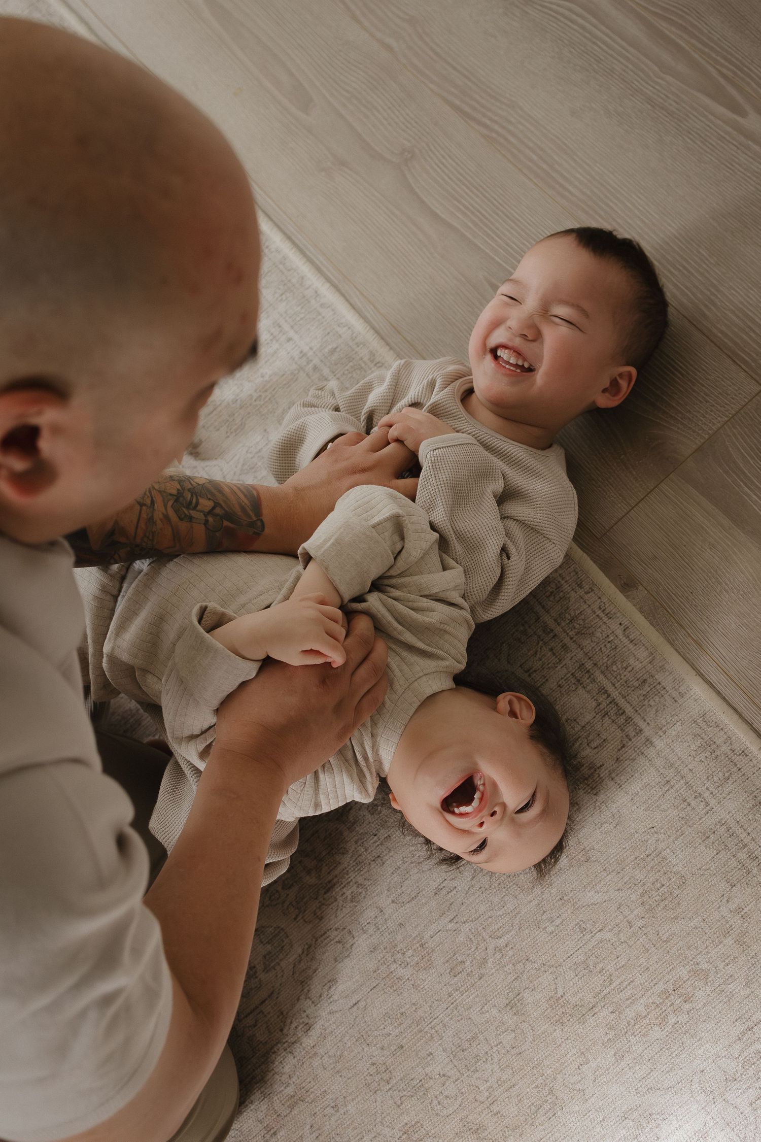 A bald tattooed parent playfully tickles two laughing babies lying on a soft rug on the floor during a lifestyle family photography session.
