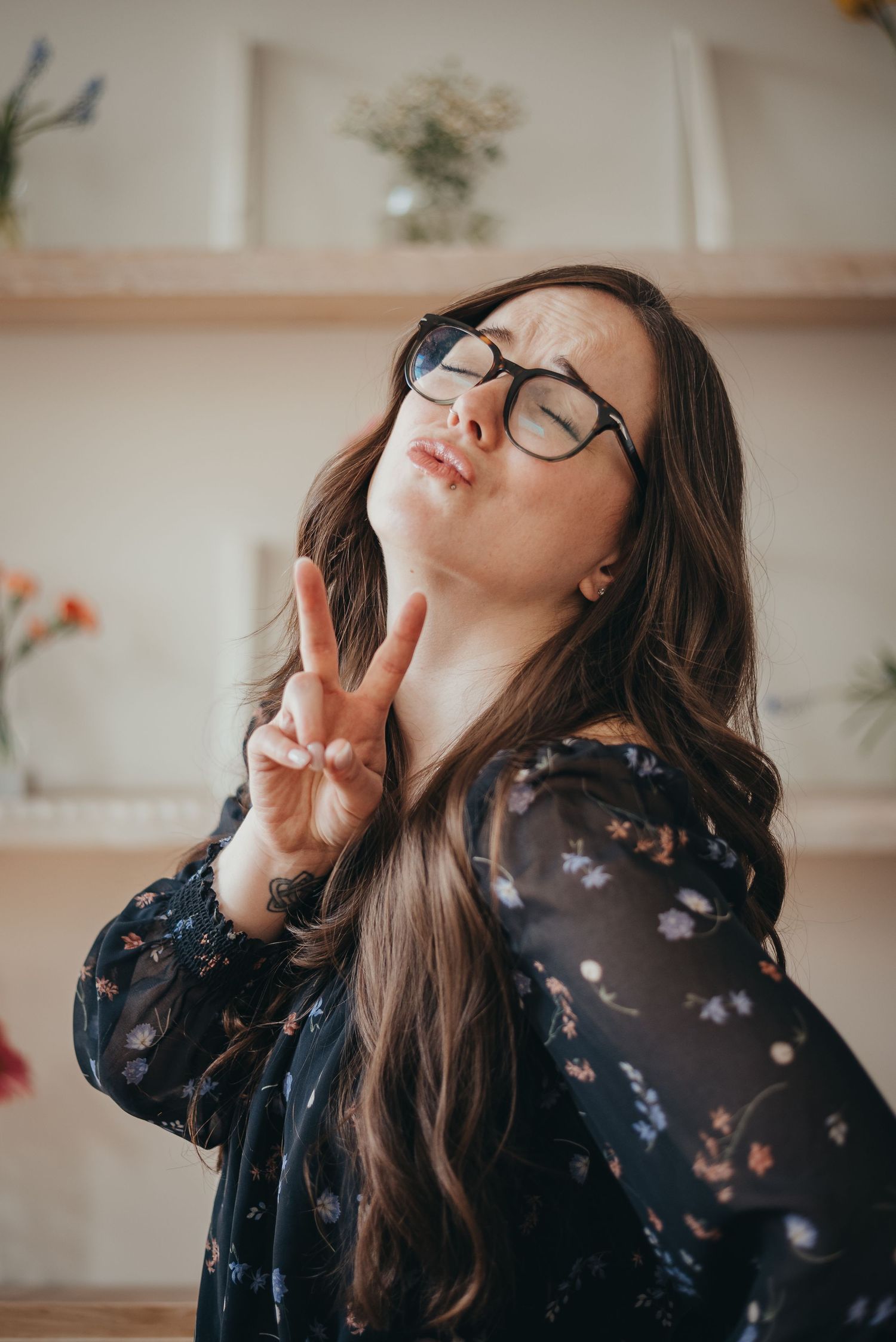 Young woman with long brown hair and glasses making a peace sign gesture, wearing a floral dress in a Calgary studio.