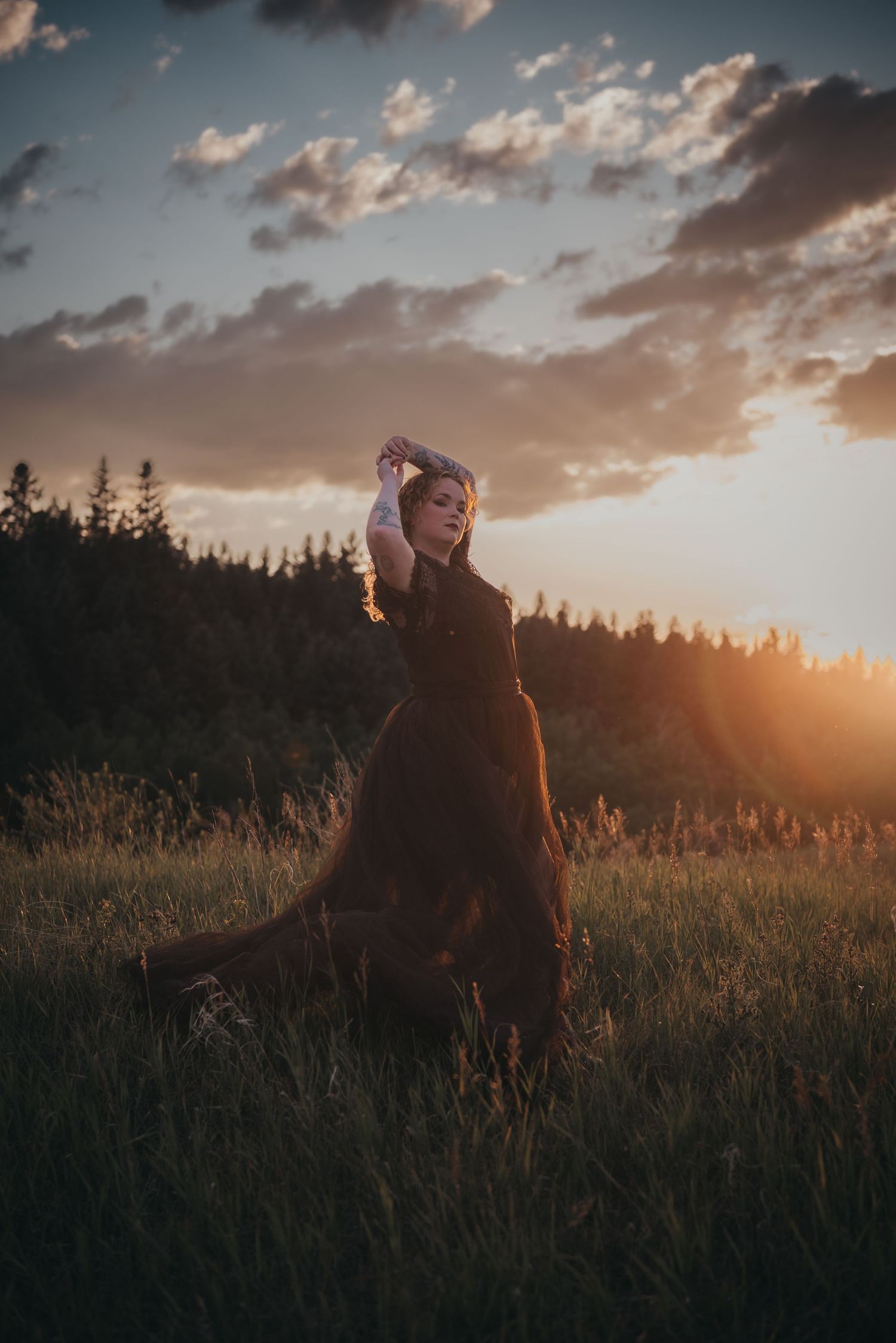 Woman in dark dress poses gracefully in golden sunset Calgary meadow with dramatic clouds and forest backdrop.