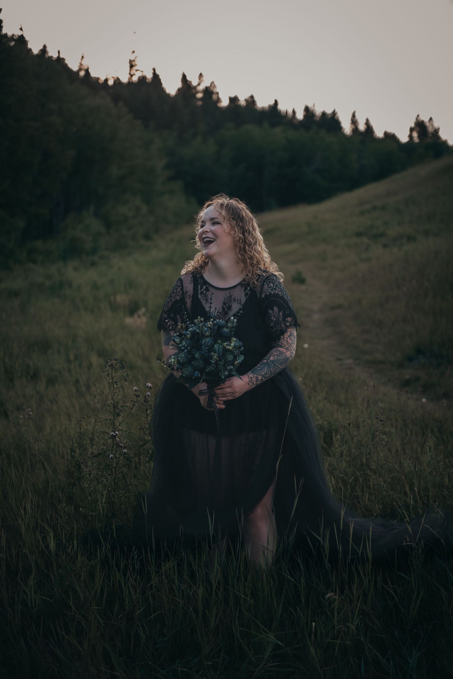 A laughing Calgary woman in a black lace gown holds a bouquet in a green meadow at dusk.