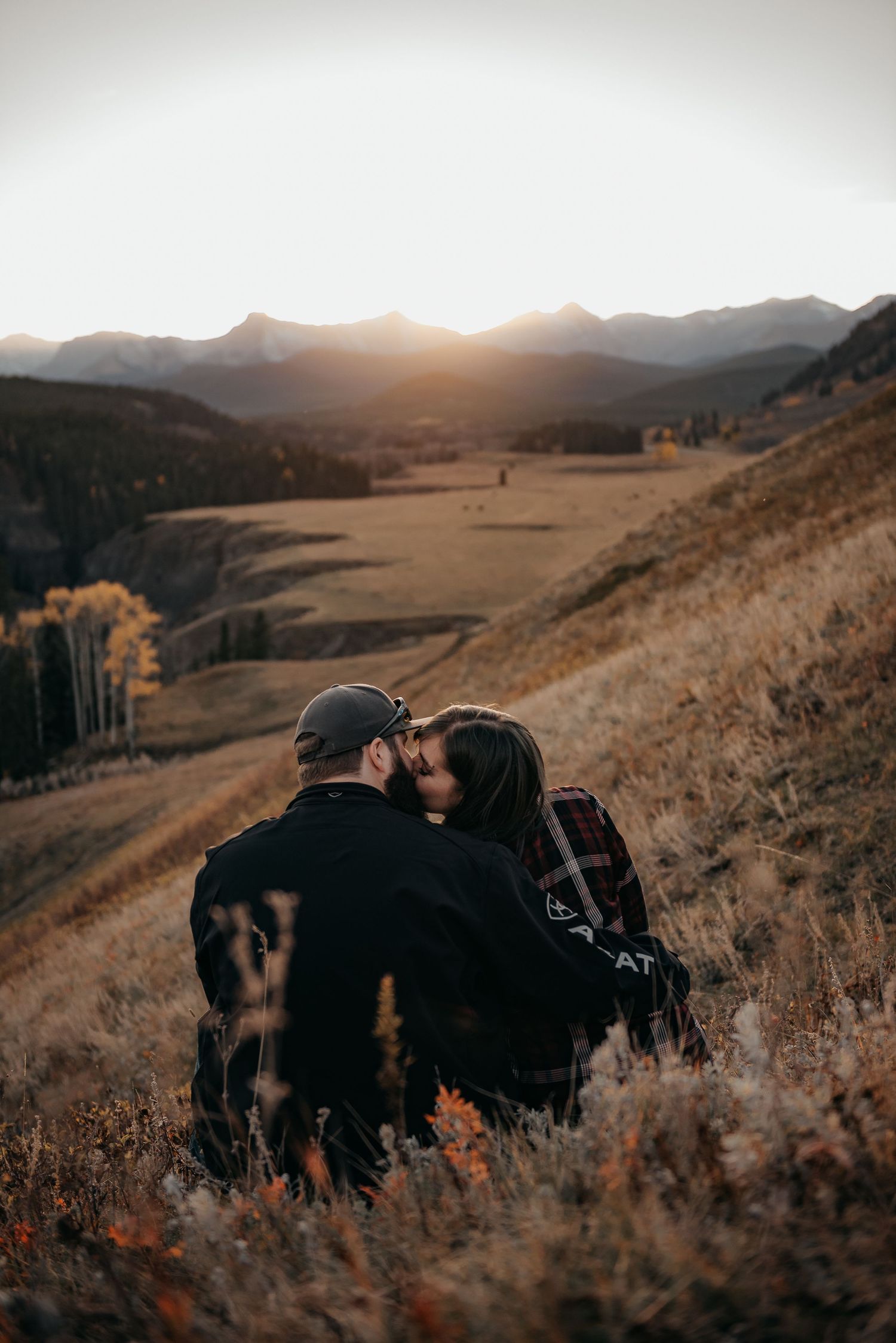 Couple sharing a romantic kiss on a golden hillside at sunset with mountain valley views in autumn.