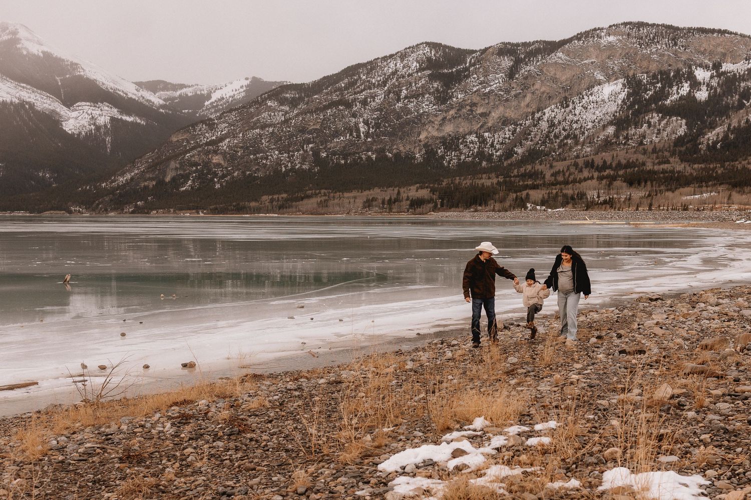 People walking along a snowy shoreline beside Barrier Lake on a winter day in a scenic landscape for a maternity session.