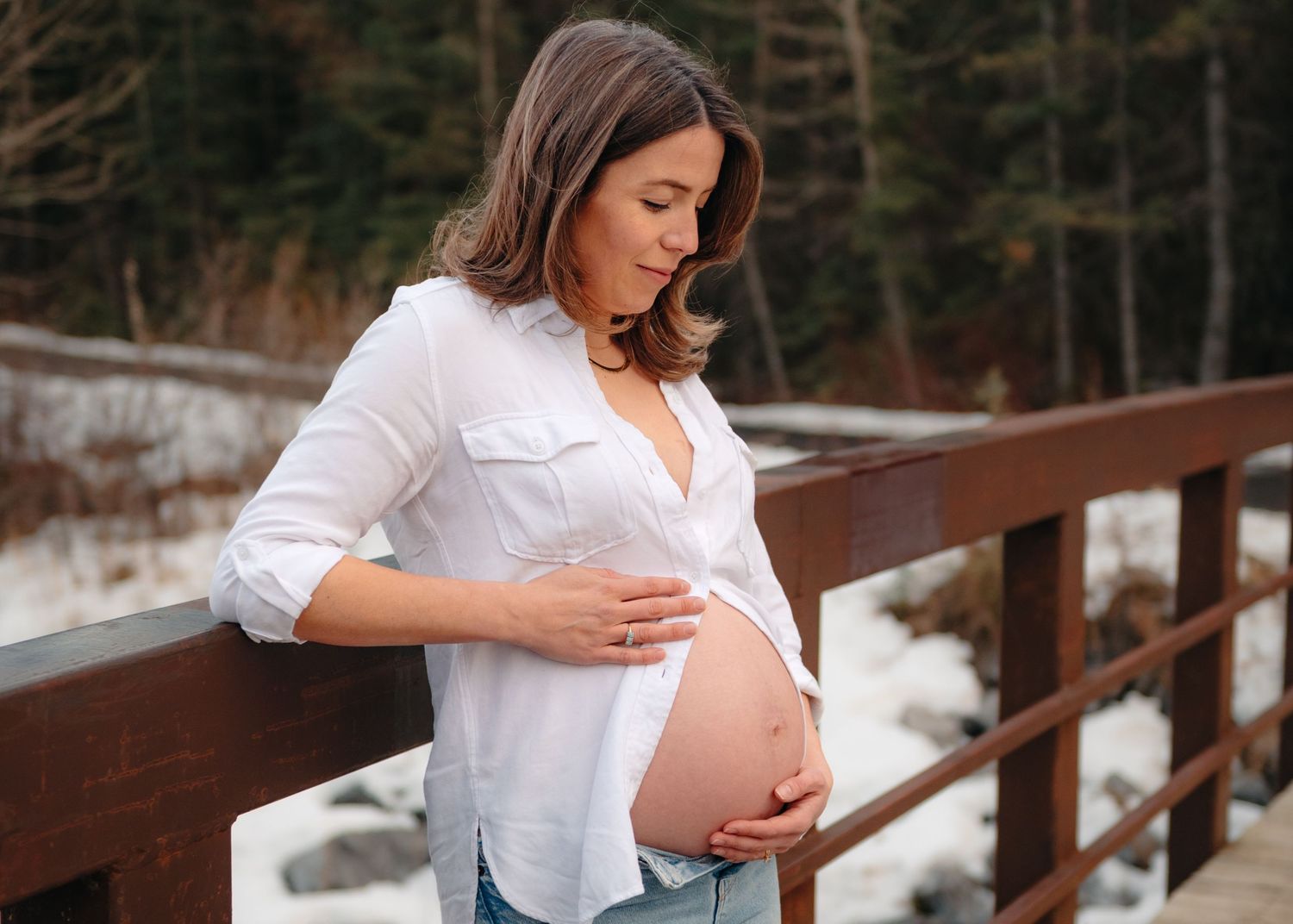 Maternity photos showing pregnant woman in white shirt and jeans on snowy wooden bridge.