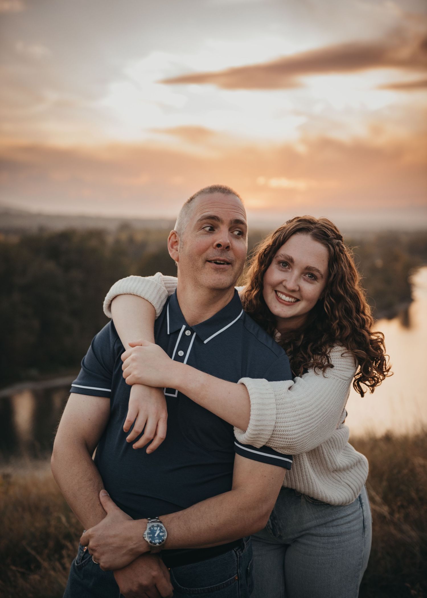 Father and daughter embracing at sunset, girl hugging man from behind, smiling outdoors with scenic Calgary sky backdrop.