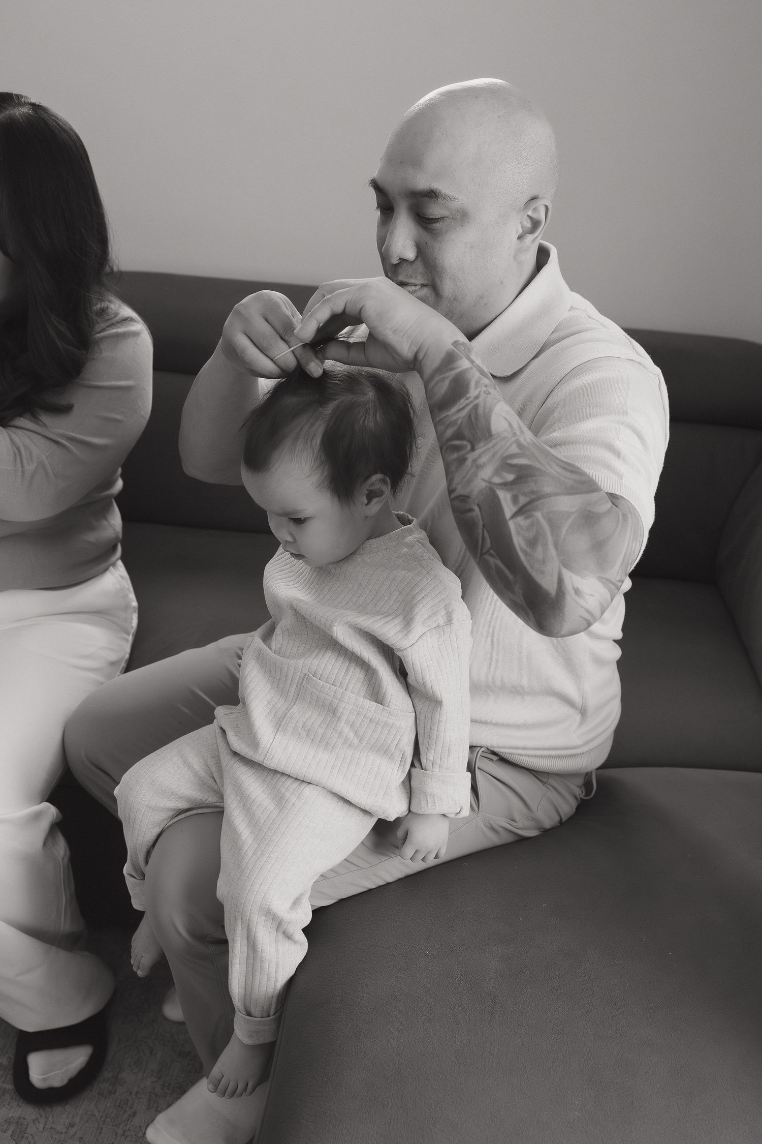 Bald father with tattoos sits on sofa styling baby's hair in black and white family lifestyle photography photo.