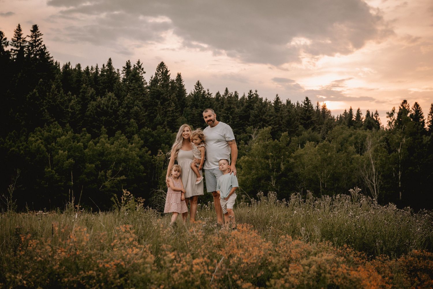Family of five posing in a golden wildflower meadow at sunset in Calgary, AB with lush green forest backdrop.