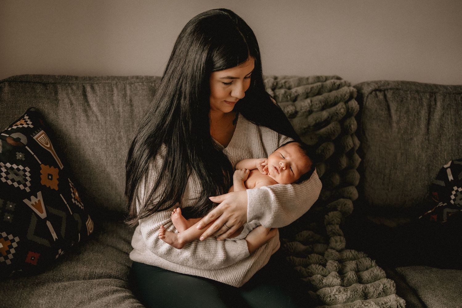 A young mother with long dark hair cradles her sleeping newborn baby on a gray couch.