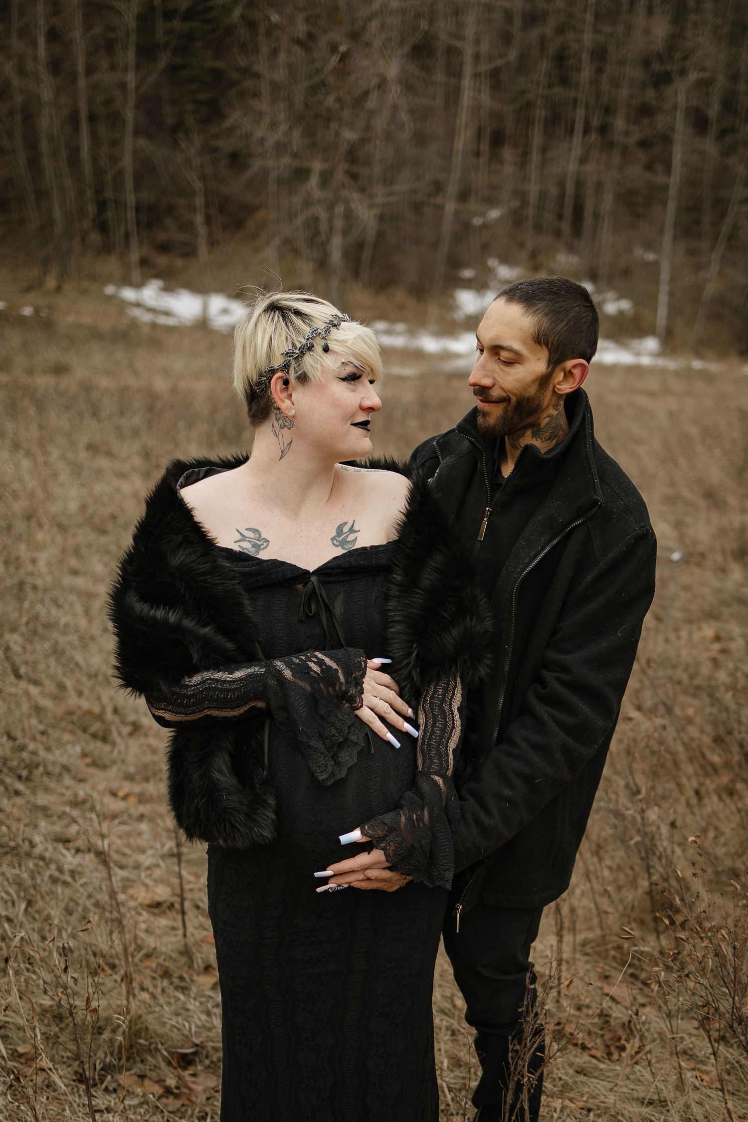 A pregnant couple in black outfits poses in a winter field, the woman wearing a fur wrap and showing tattoos.