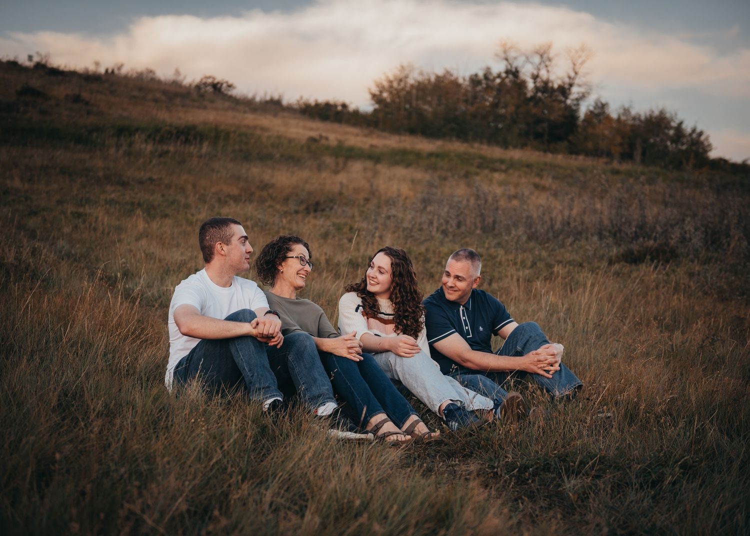 Four family members sitting together laughing in a golden grass field at dusk, enjoying a candid outdoor portrait session.