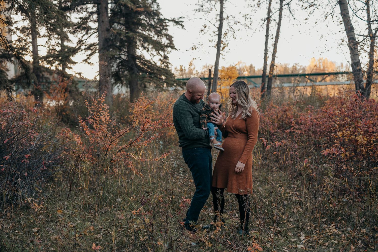 Family of three posing in Bowness Park, parents holding toddler, golden fall foliage and trees in background.