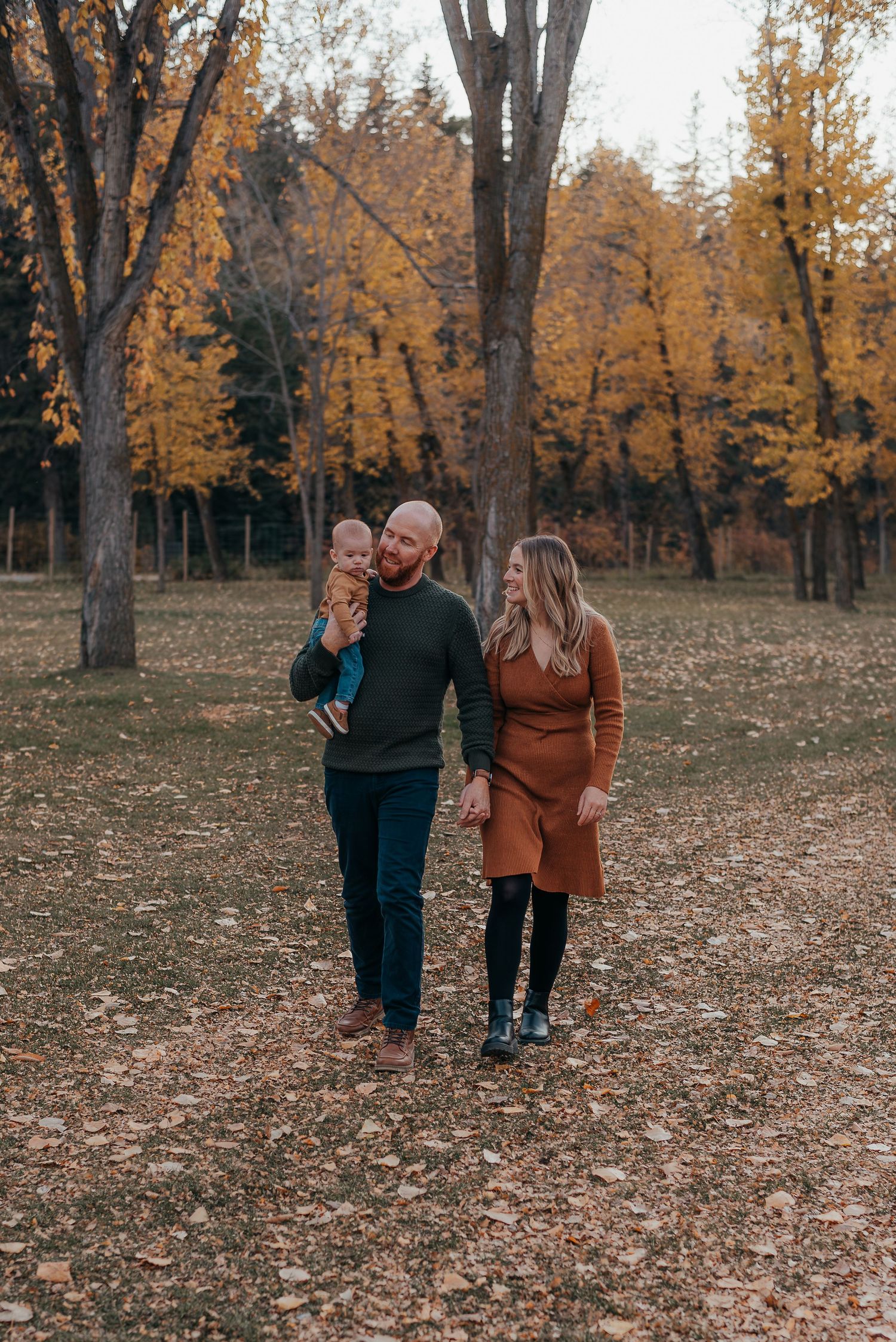 A family of three walks through Bowness Park, dad holding baby, mom in rust dress, surrounded by golden fall trees for Calgary photographer