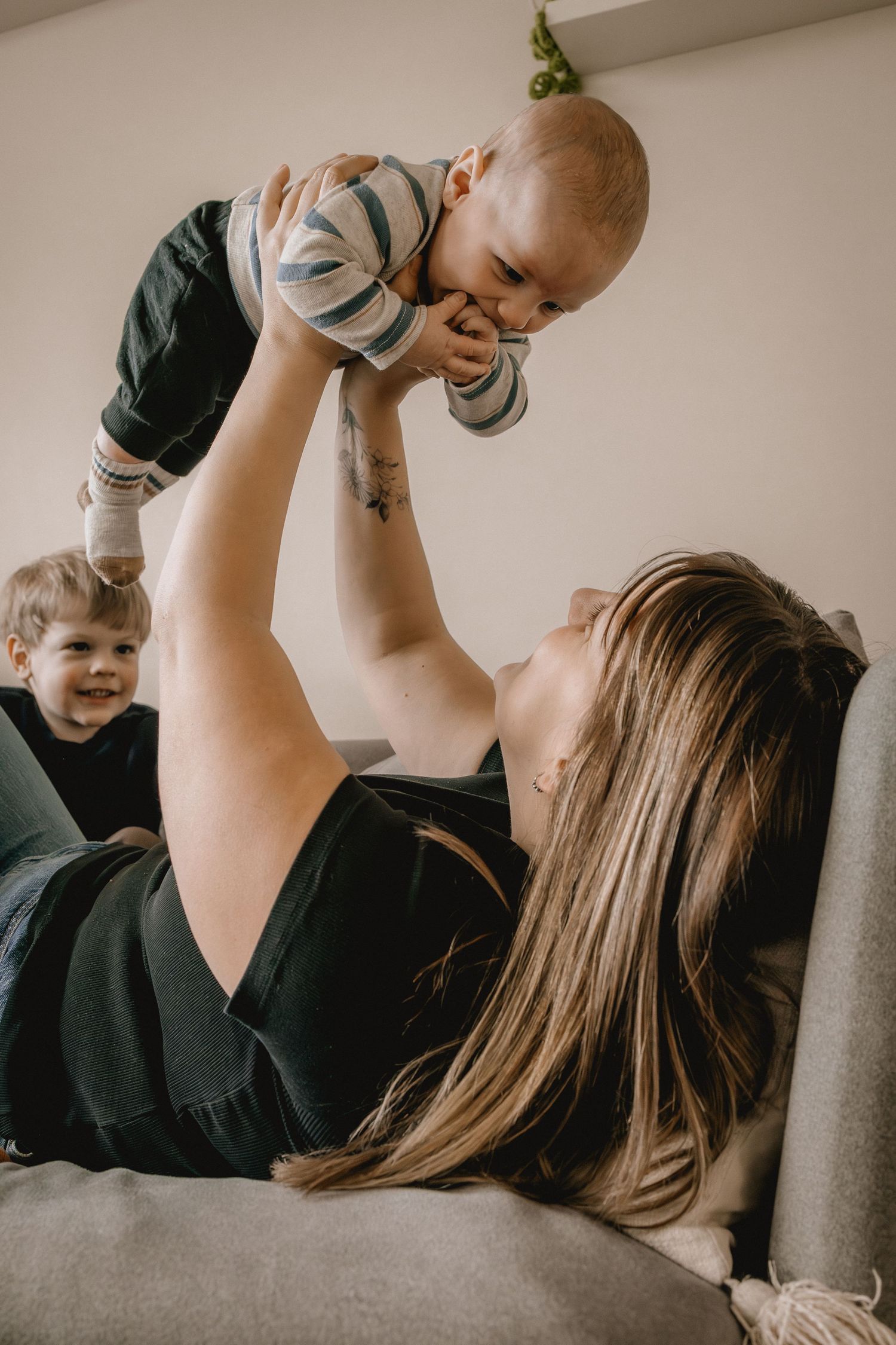 A mother lying on a couch joyfully lifts her baby in the air while her toddler smiles beside her for a Calgary photography session