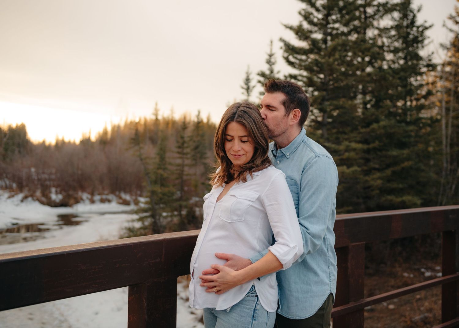 Couple sharing a tender moment on a Calgary walking bridge at sunset, with the woman's pregnant belly cradled gently outdoors.