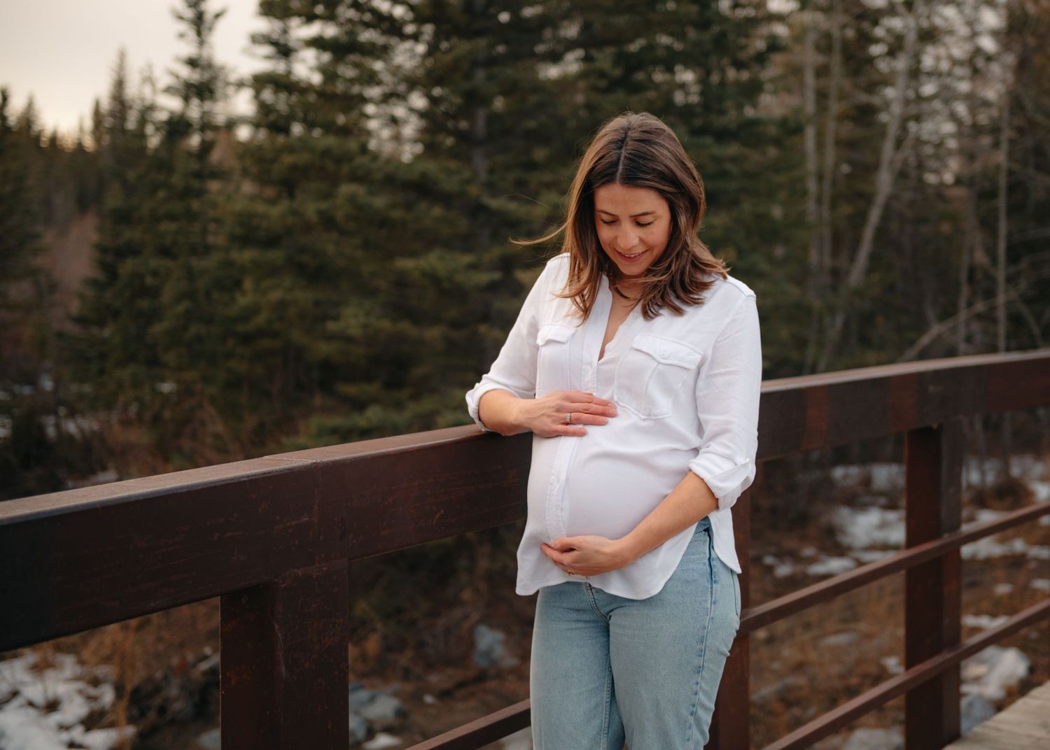 Winter maternity photos on bridge surrounded by snow-covered evergreen trees in Calgary, AB.