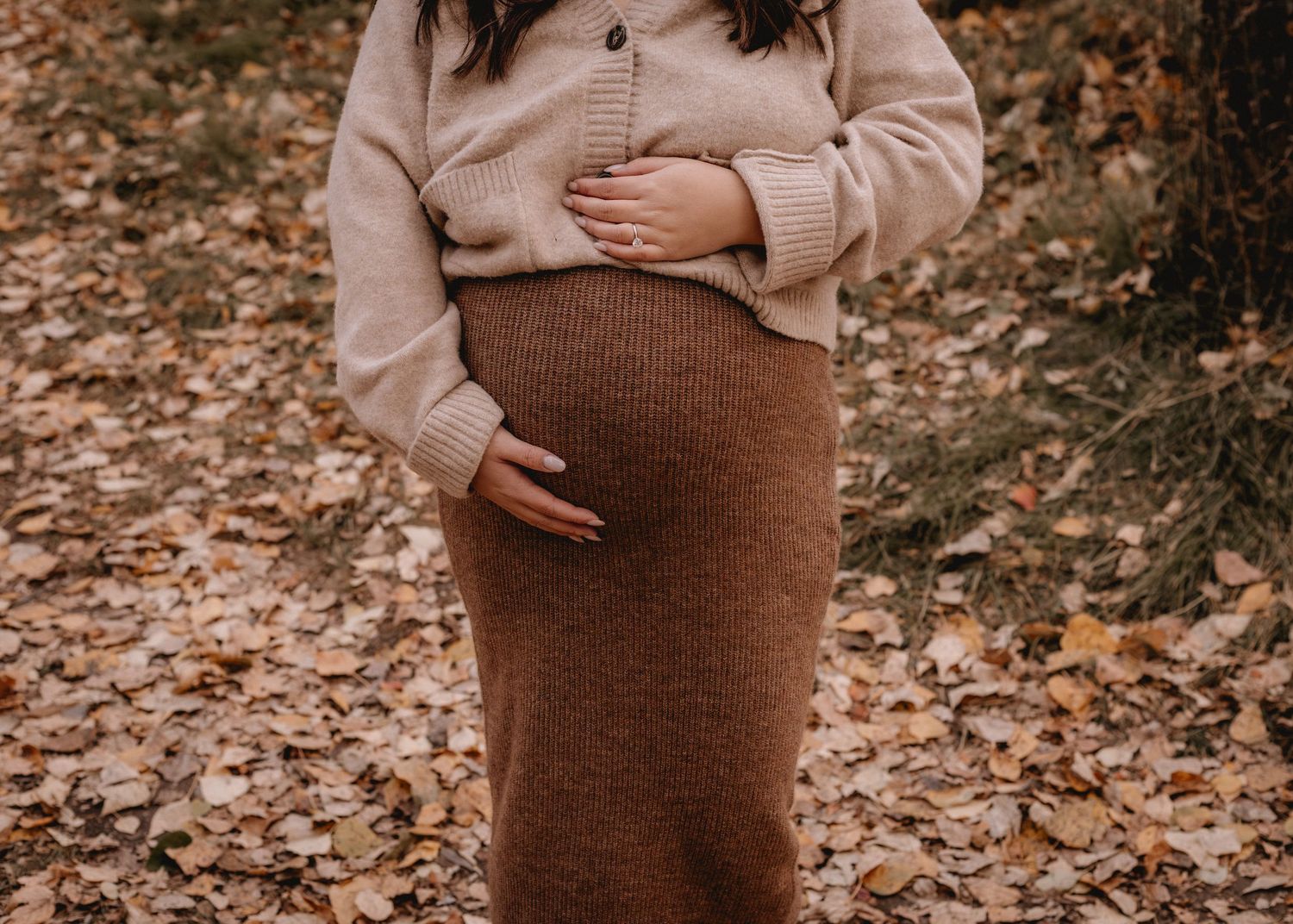 Woman in brown knit dress cradling her baby bump in an autumn setting.