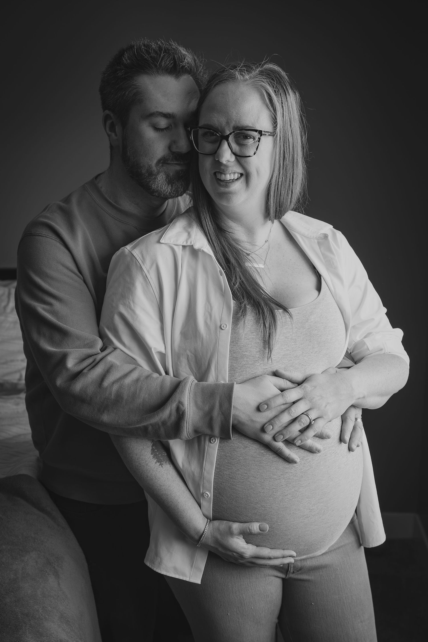 Black and white photo of a smiling pregnant woman with glasses, embraced from behind by her partner, both cradling her baby bump.