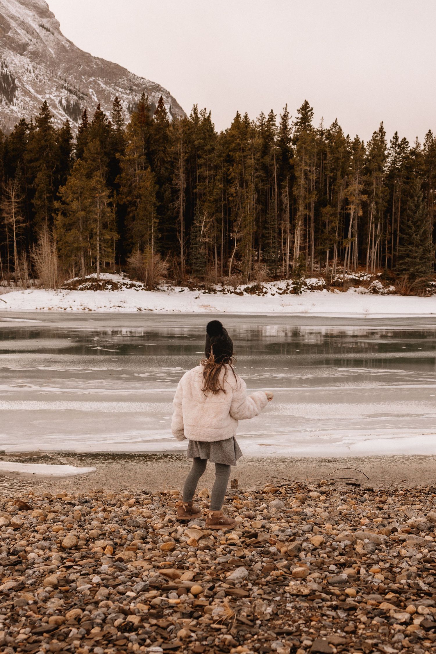 Girl in white fur coat standing on rocky shore of partially frozen Barrier Lake, AB surrounded by snowy pine forest.
