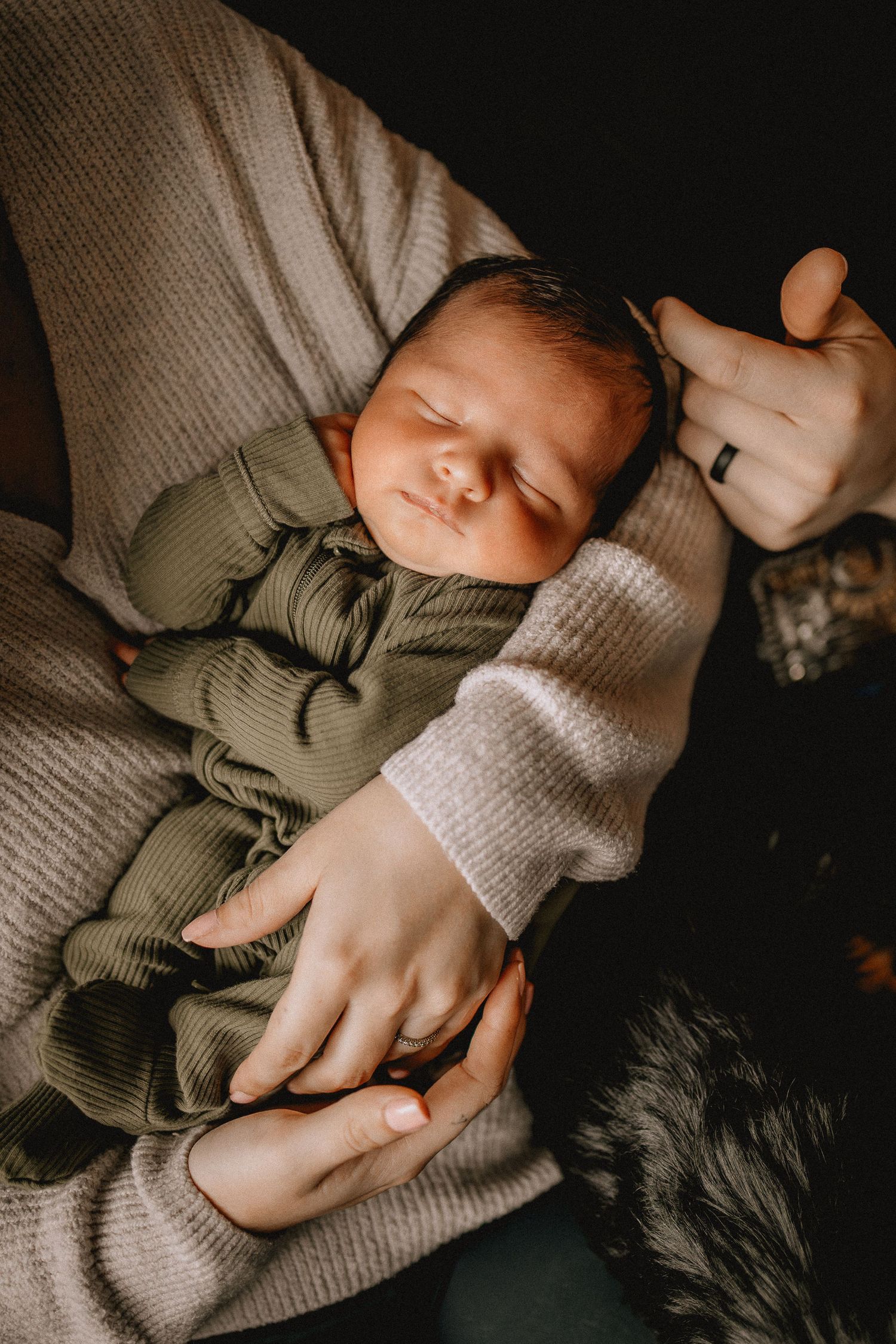 Newborn baby sleeping peacefully in olive green onesie, cradled in parent's arms wearing a beige knit sweater.