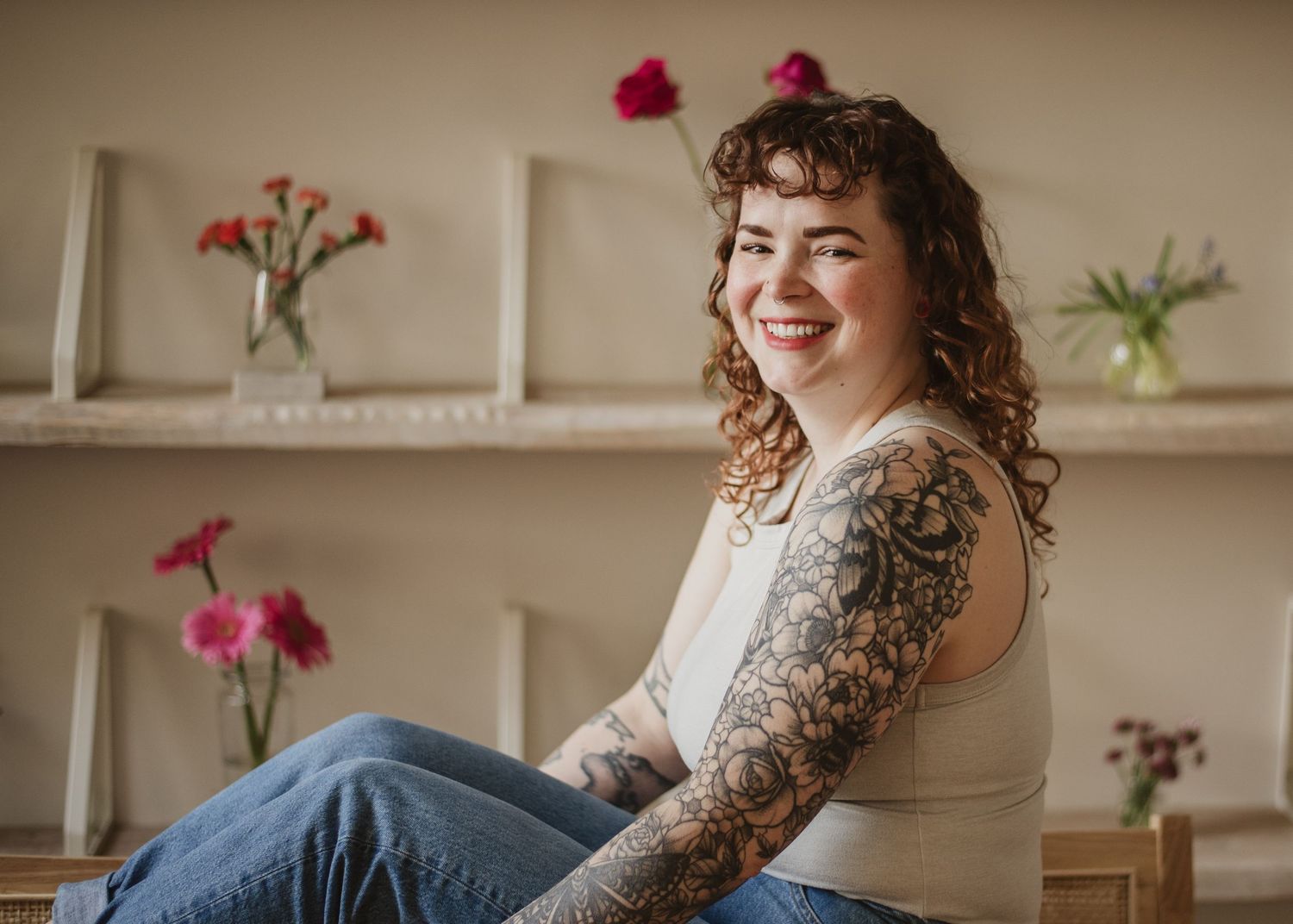 Smiling woman with tattoo sleeve sitting casually in jeans and white top, pink flowers on shelves behind her.