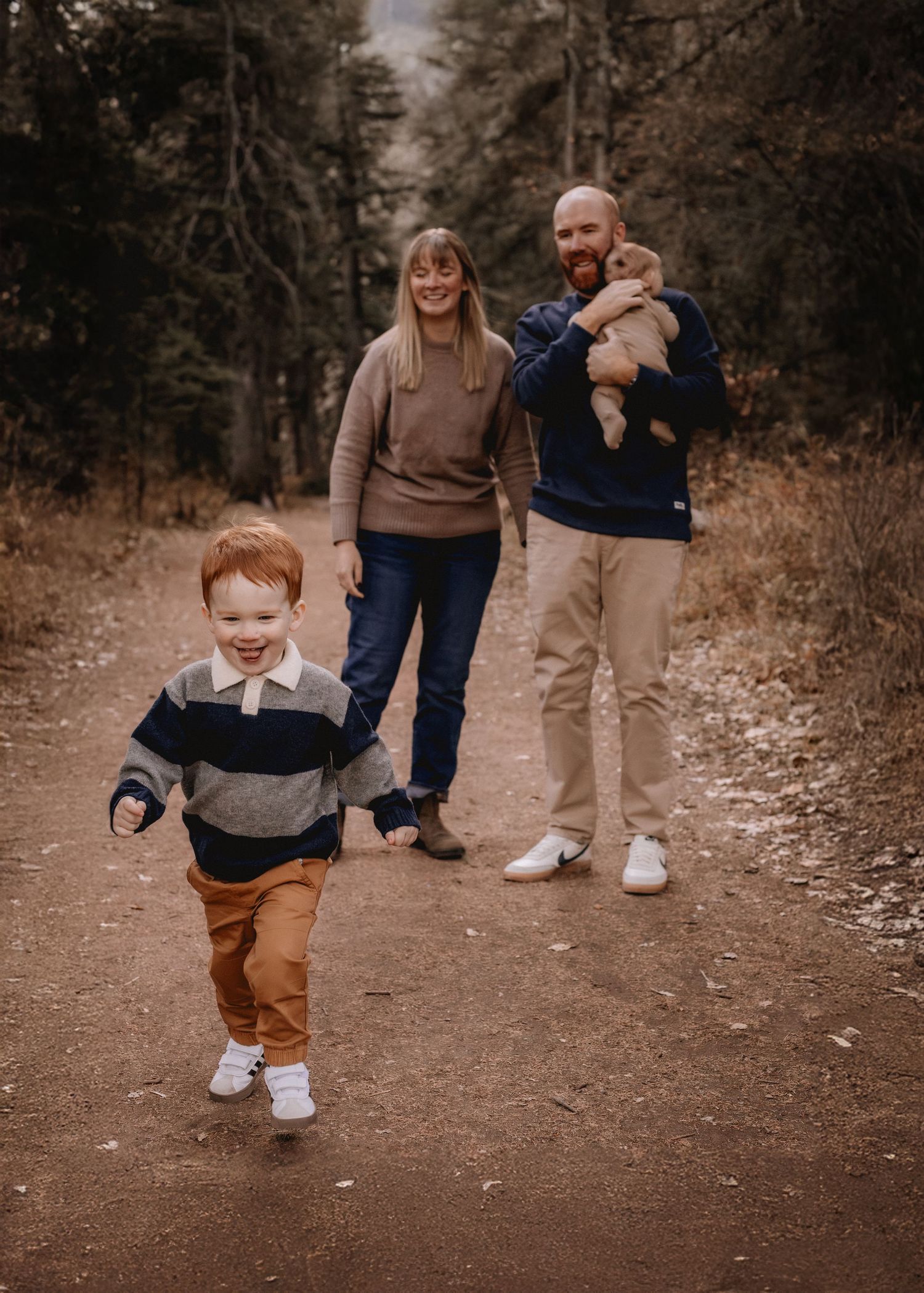 Family of four posing on a Calgary forest trail, young boy running toward camera, parents smiling behind him.
