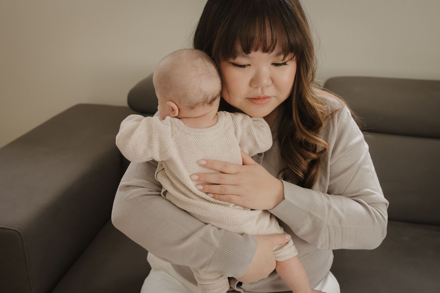 An in-home Calgary, AB lifestyle session of a young mother tenderly holding her newborn baby against her chest while sitting on a gray sofa.