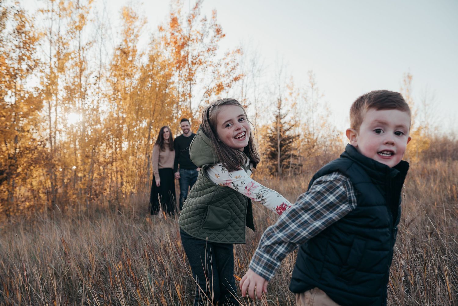 Family photo session in golden autumn field, smiling girl reaching for playful toddler boy, parents standing in background.