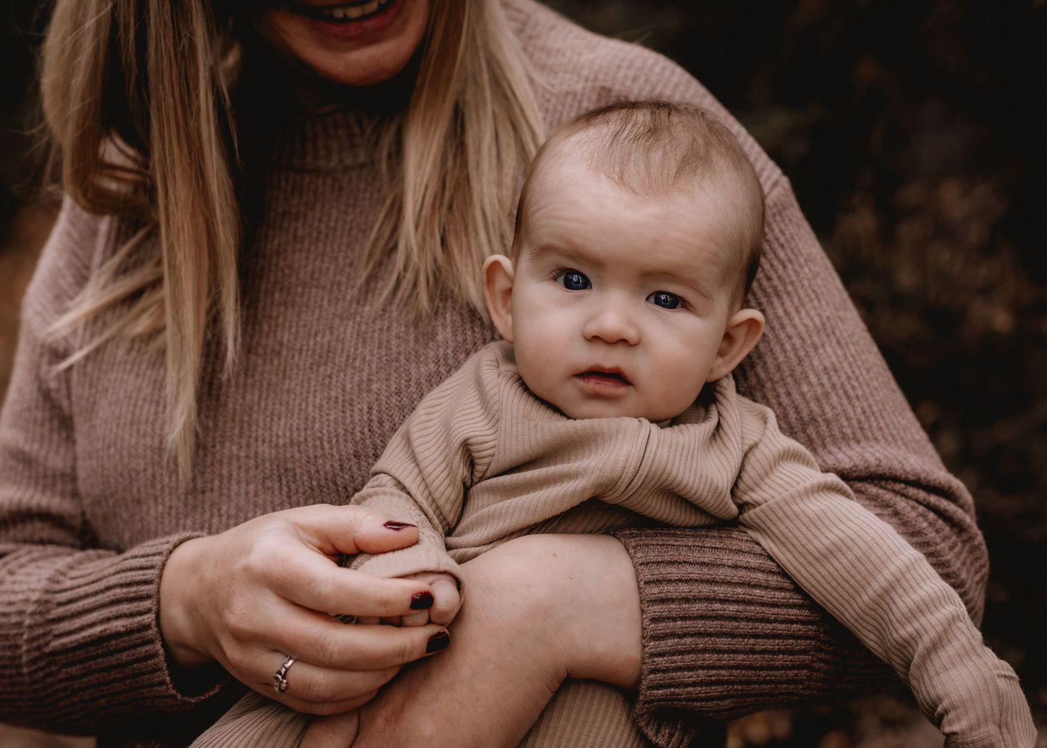 A mother in a beige knit sweater tenderly holds her curious baby dressed in a neutral onesie in Fish Creek Park.