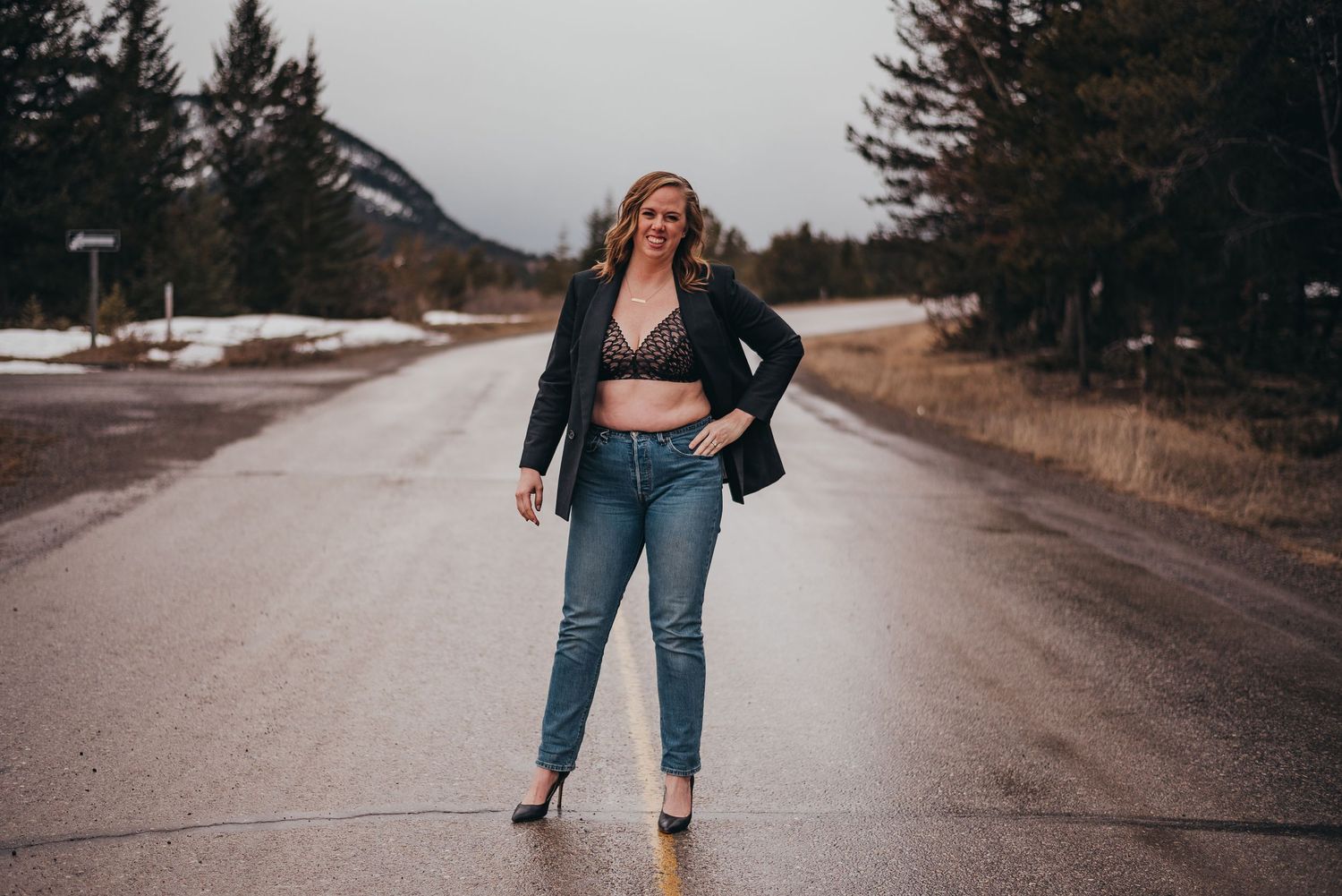 Woman in black blazer and jeans poses confidently on a mountain road with snowy landscape background.