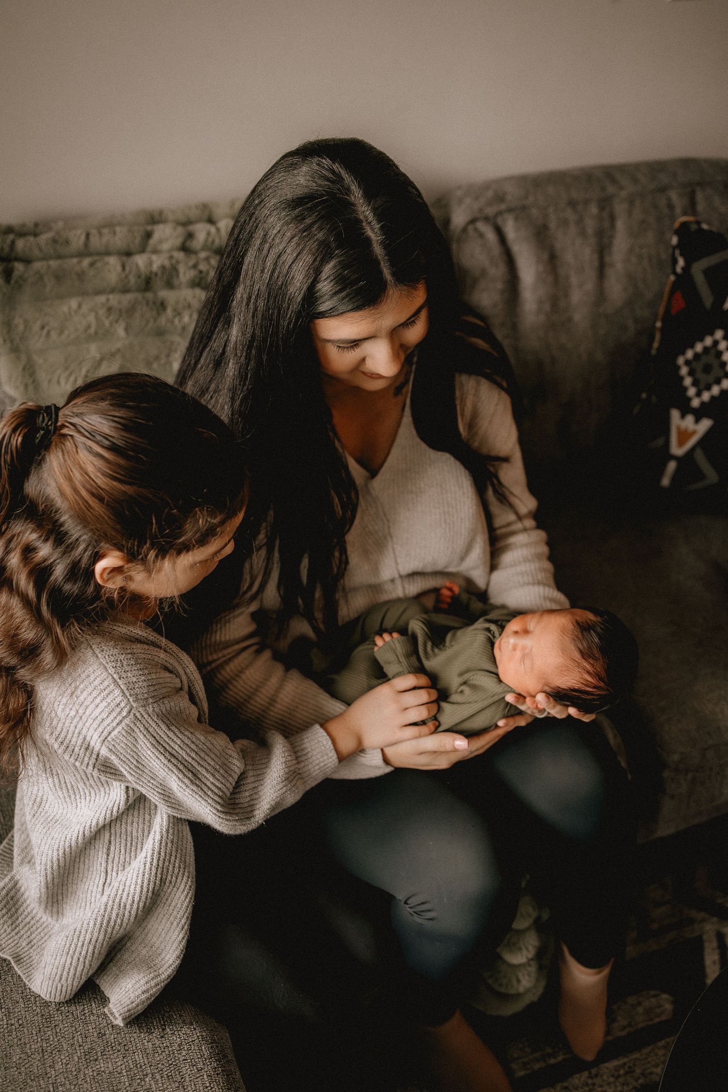 A Calgary mother and young girl sit on a couch, lovingly gazing at a newborn baby wrapped in green.