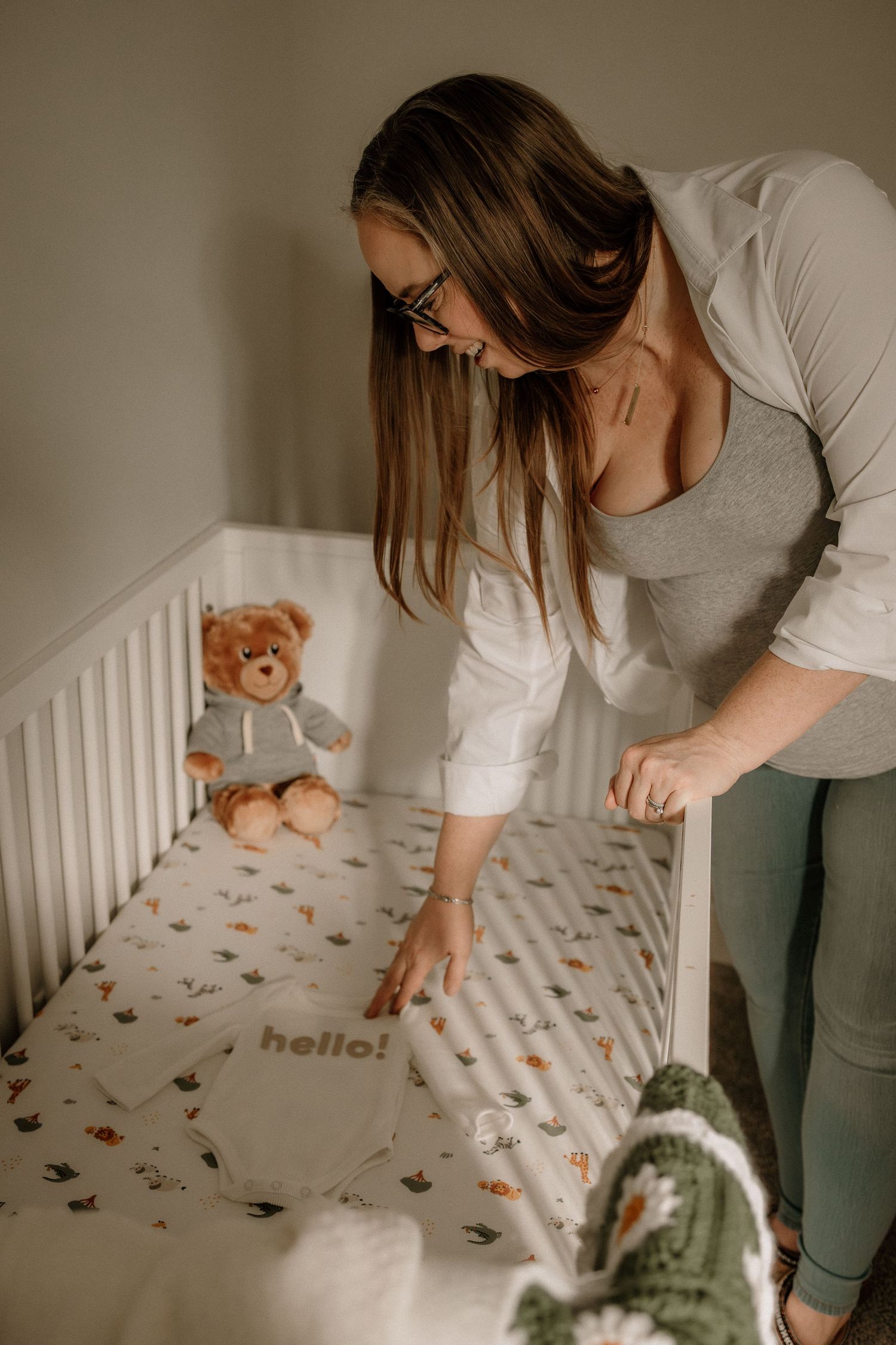 Maternity session of a woman arranging a hello onesie in a white crib with a teddy bear on patterned sheet in Calgary, AB.