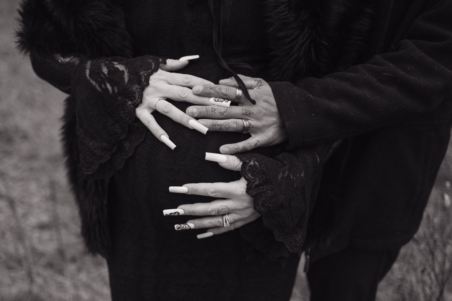 Black and white photo of two hands with long acrylic nails and rings clasped together over a dark outfit.