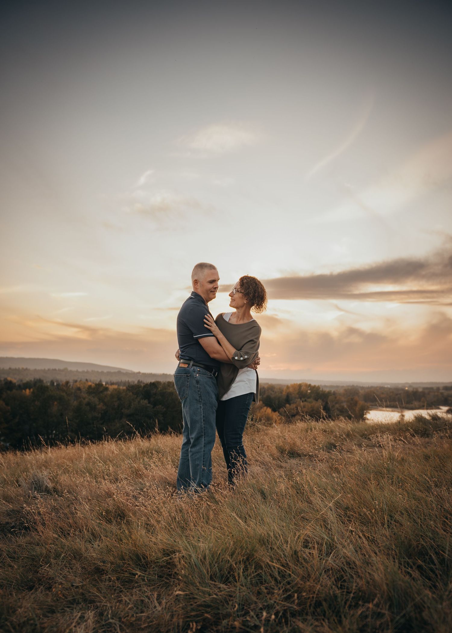 Calgary couple embracing at golden hour on a grassy hilltop with a scenic sunset sky and distant landscape behind them.